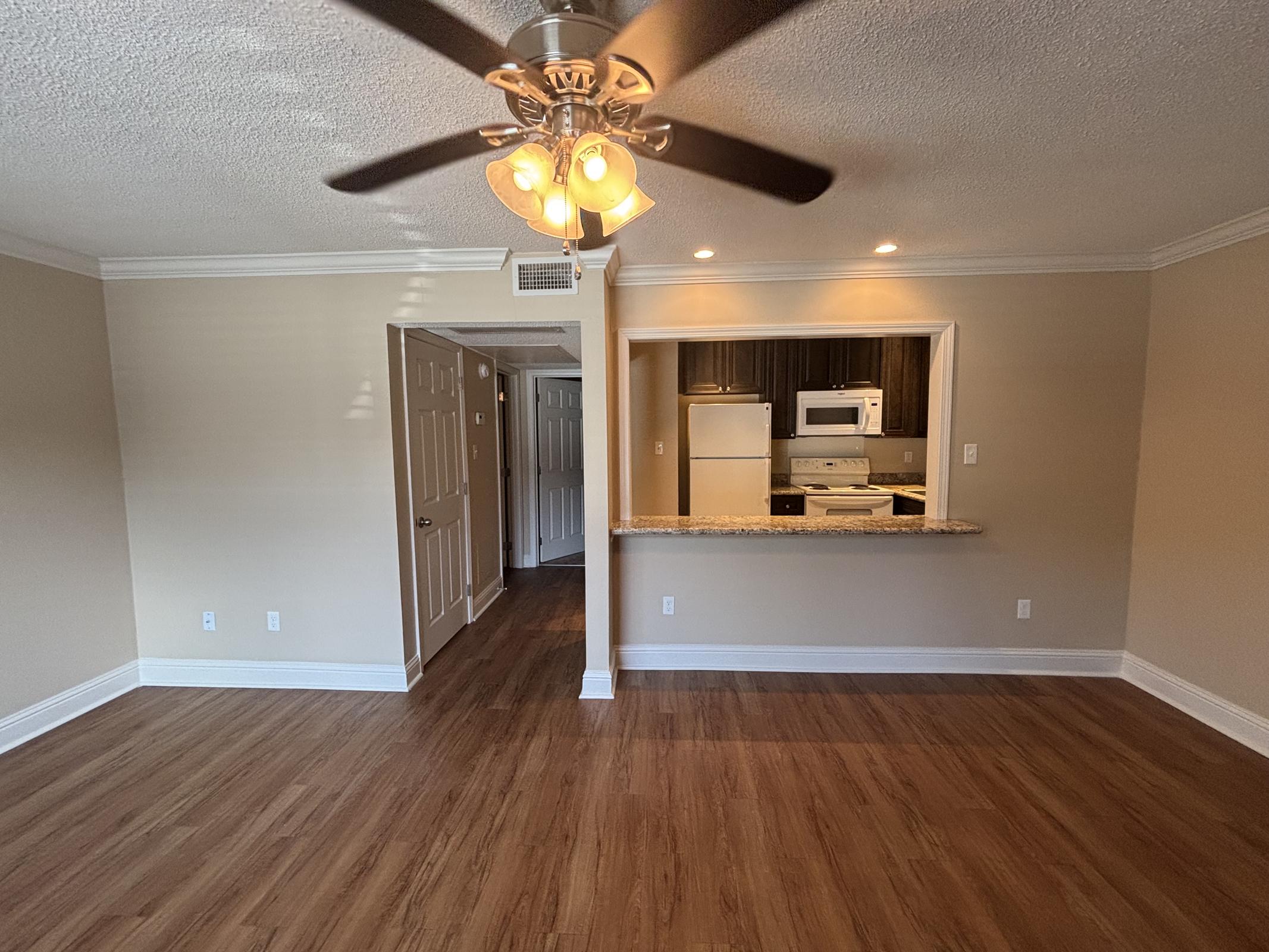 A spacious living area featuring a ceiling fan, light-colored walls, and wooden flooring. A doorway leads to another room, while an open kitchen with dark cabinets and a white refrigerator is visible in the background. Natural light brightens the space, creating a warm and inviting atmosphere.