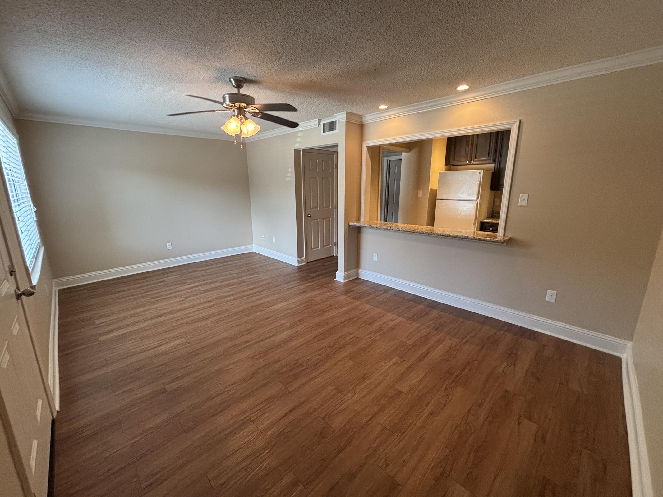 A spacious, empty living room with light brown wooden flooring. A ceiling fan hangs from the textured ceiling. There is a large window with blinds, and an open wall leads to a kitchen area with a bar counter. The walls are painted in a neutral color, creating a bright and airy atmosphere.