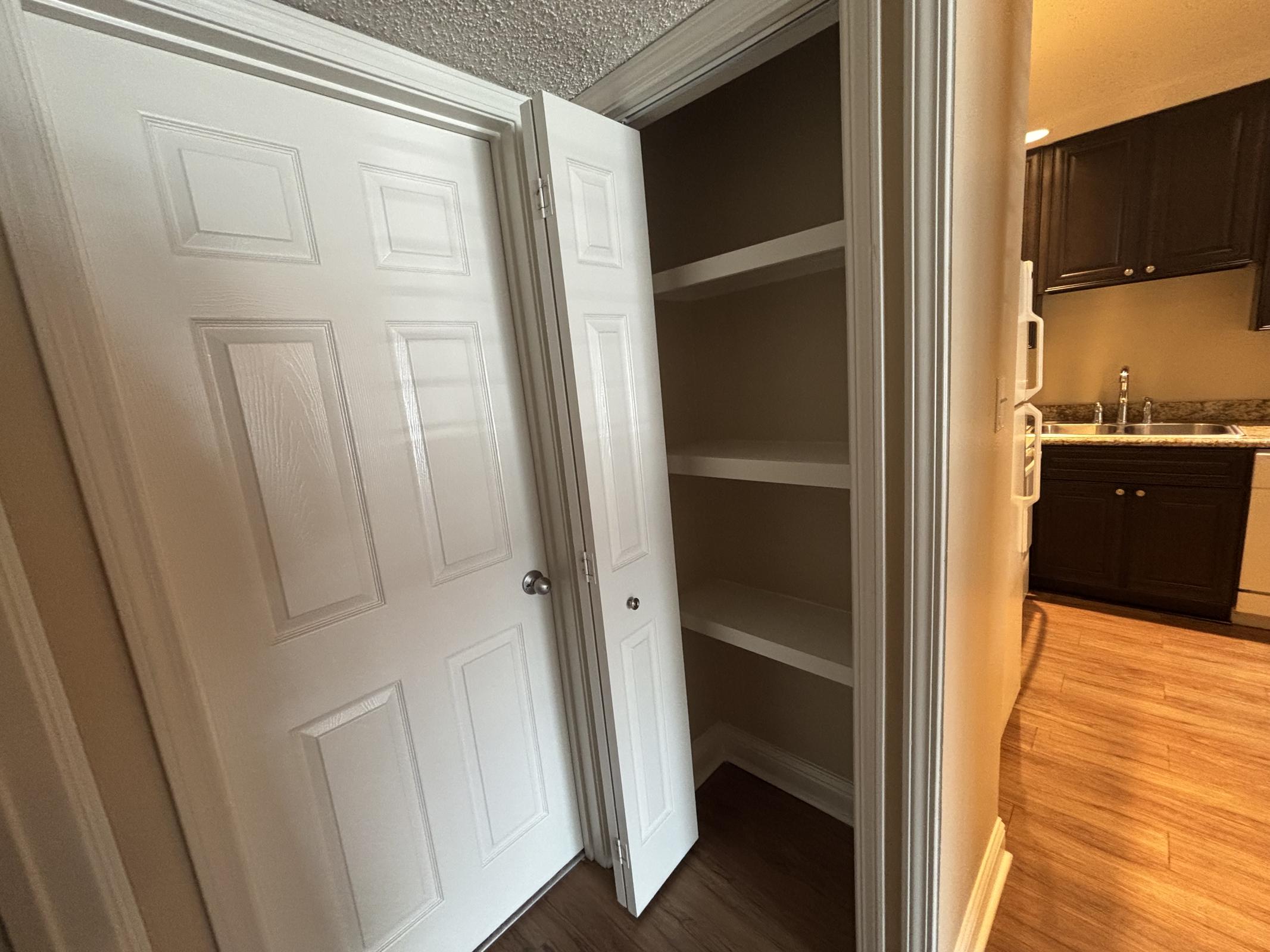 A view of a narrow pantry or storage closet with two white doors partially open, revealing empty shelves inside. The doorway leads to a kitchen area with dark cabinetry and a sink visible on the right. The floor has a warm wood finish, and the walls are painted a light neutral color.