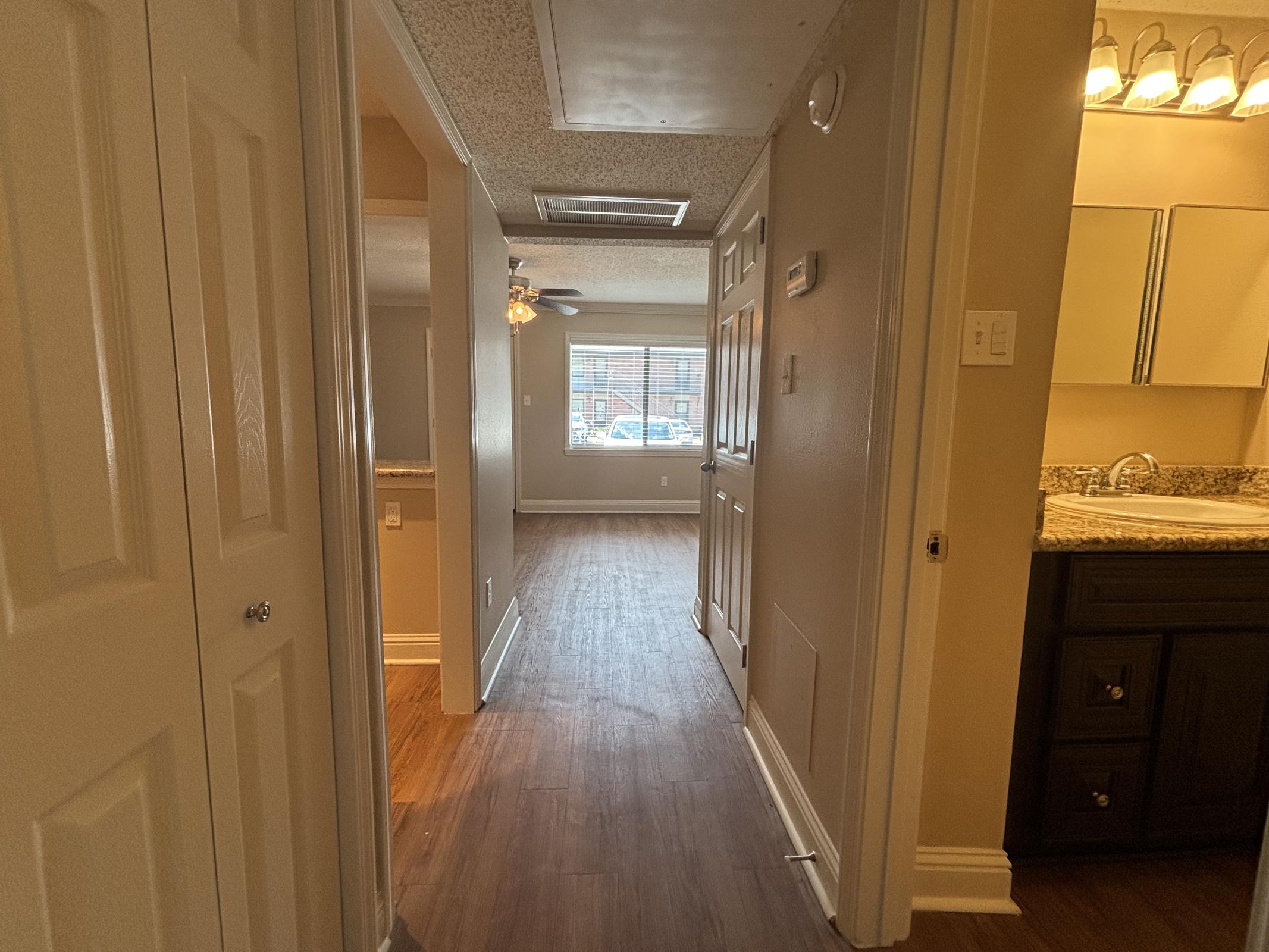 View of a hallway leading into a living space with wood-style flooring. The hallway is flanked by beige walls and has a door on the left. At the end, there’s a window providing natural light. To the right, a partially visible kitchen area with dark cabinetry and a granite countertop is seen.