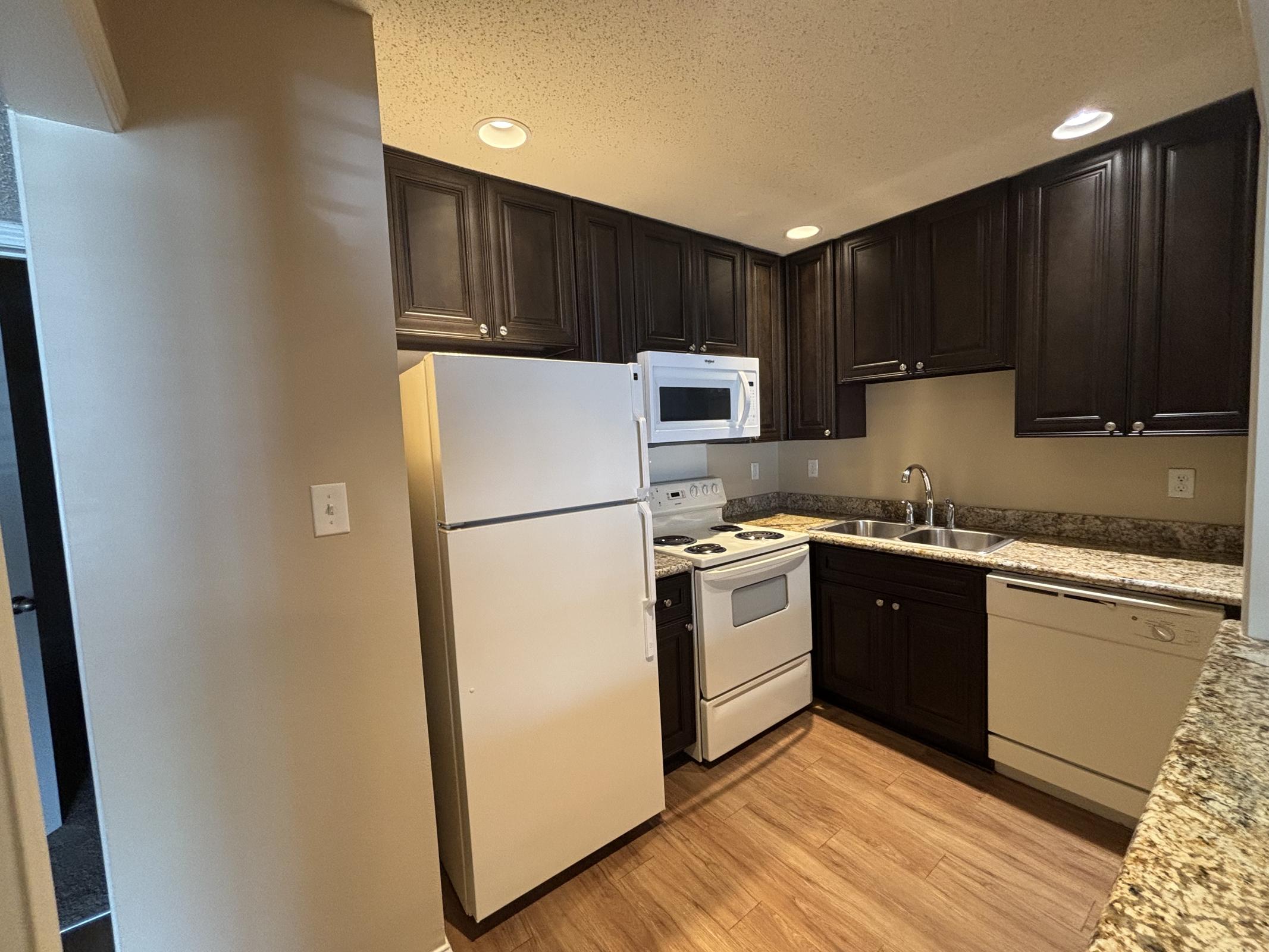 A modern kitchen featuring dark wood cabinets, granite countertops, and stainless steel appliances. The layout includes a refrigerator, stove, microwave, and dishwasher. The flooring is hardwood, and there are recessed lights in the ceiling. The space appears clean and well-maintained.