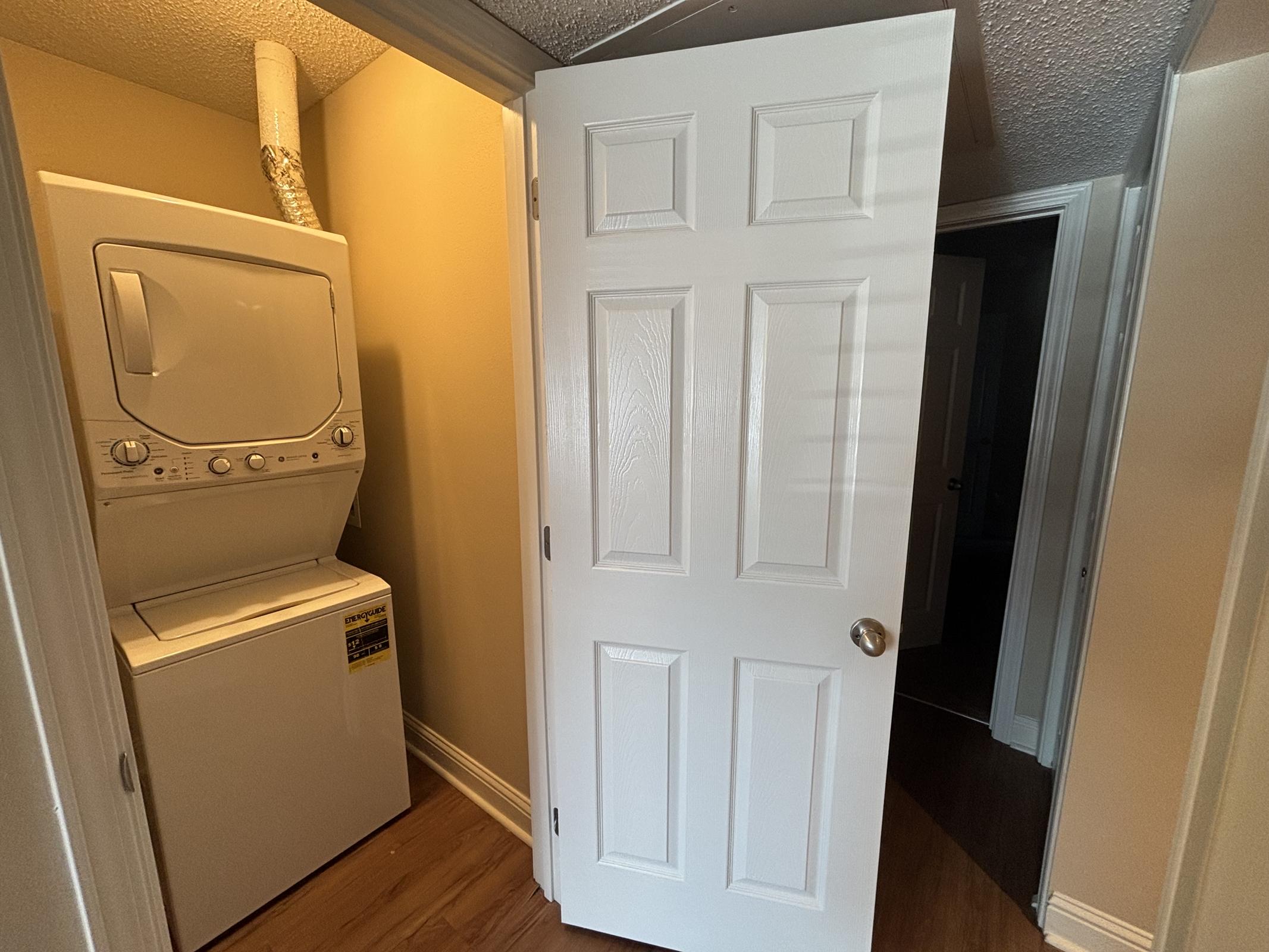 A white door partially open, leading to a laundry area with a stacked washer and dryer. The walls are painted in a warm hue, and the floor is wooden. A hallway is visible beyond the door, suggesting access to other rooms.