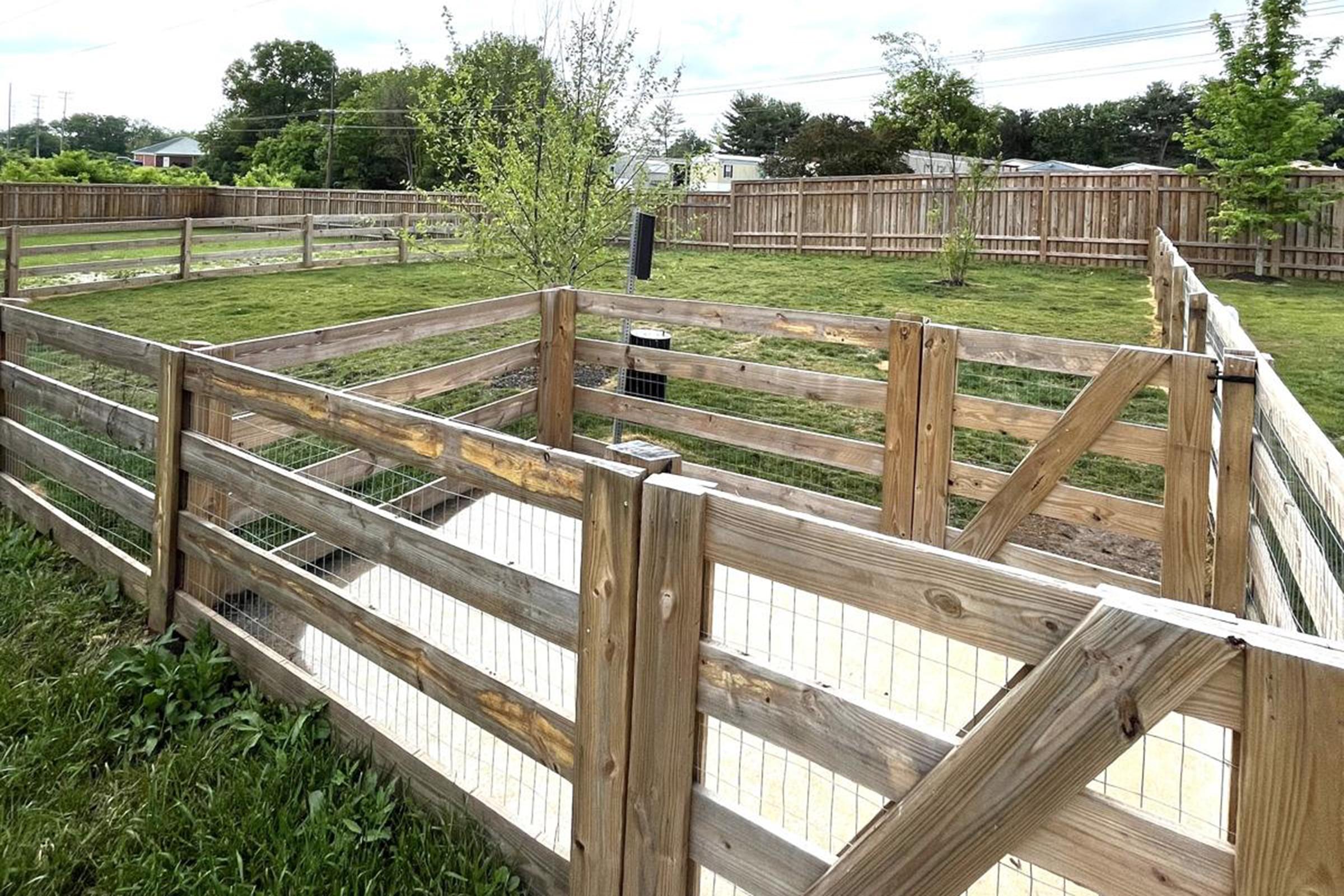 A wooden fenced area in a grassy field, featuring multiple sections and gates. Small trees or shrubs are planted within the enclosure, and the landscape in the background includes more fenced areas and distant structures under a cloudy sky.