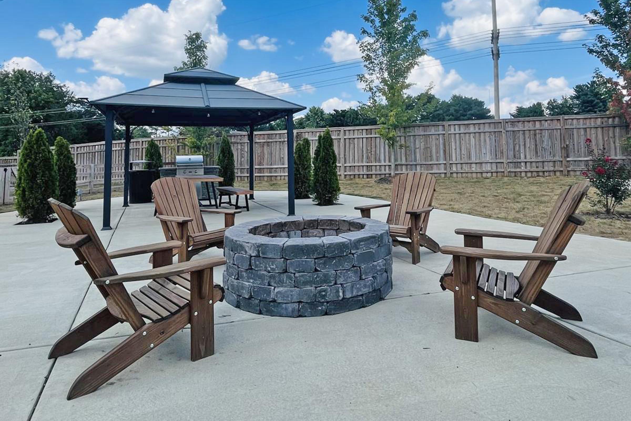 A cozy outdoor space featuring a stone fire pit surrounded by wooden Adirondack chairs. In the background, there's a gazebo and a grass area with young trees, under a bright blue sky with fluffy clouds. The setting is ideal for gatherings or relaxation.