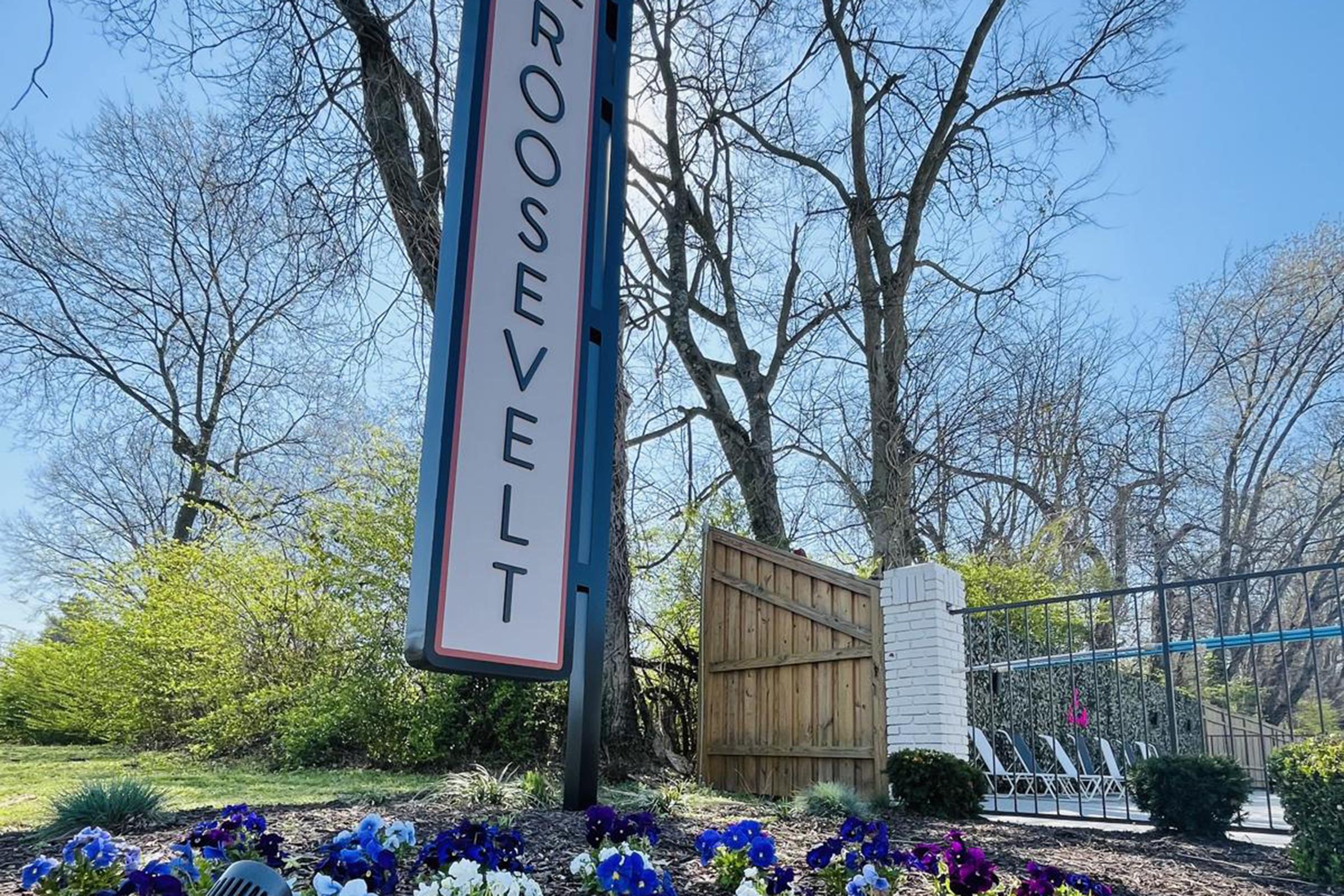 A sign reading "Roosevelt" stands prominently in a landscaped area. Surrounding the sign are vibrant flowers in shades of purple and white, with green bushes in the background. Leafless trees are visible against a clear blue sky, indicating early spring or late winter. A wooden fence is partially visible behind the sign.