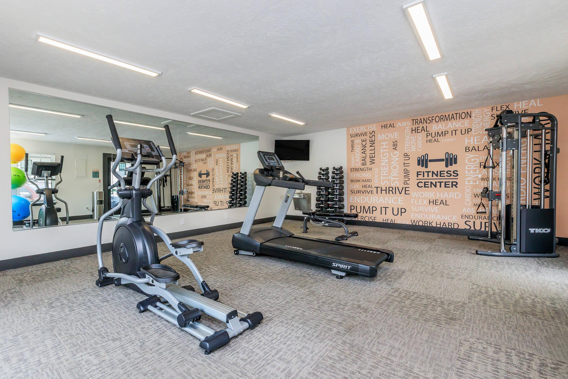 A modern fitness center featuring an elliptical machine, a treadmill, and a multi-use weight machine. The walls are decorated with motivational fitness phrases, and large mirrors are present. Various weights are neatly arranged on a rack, creating an inviting workout environment.