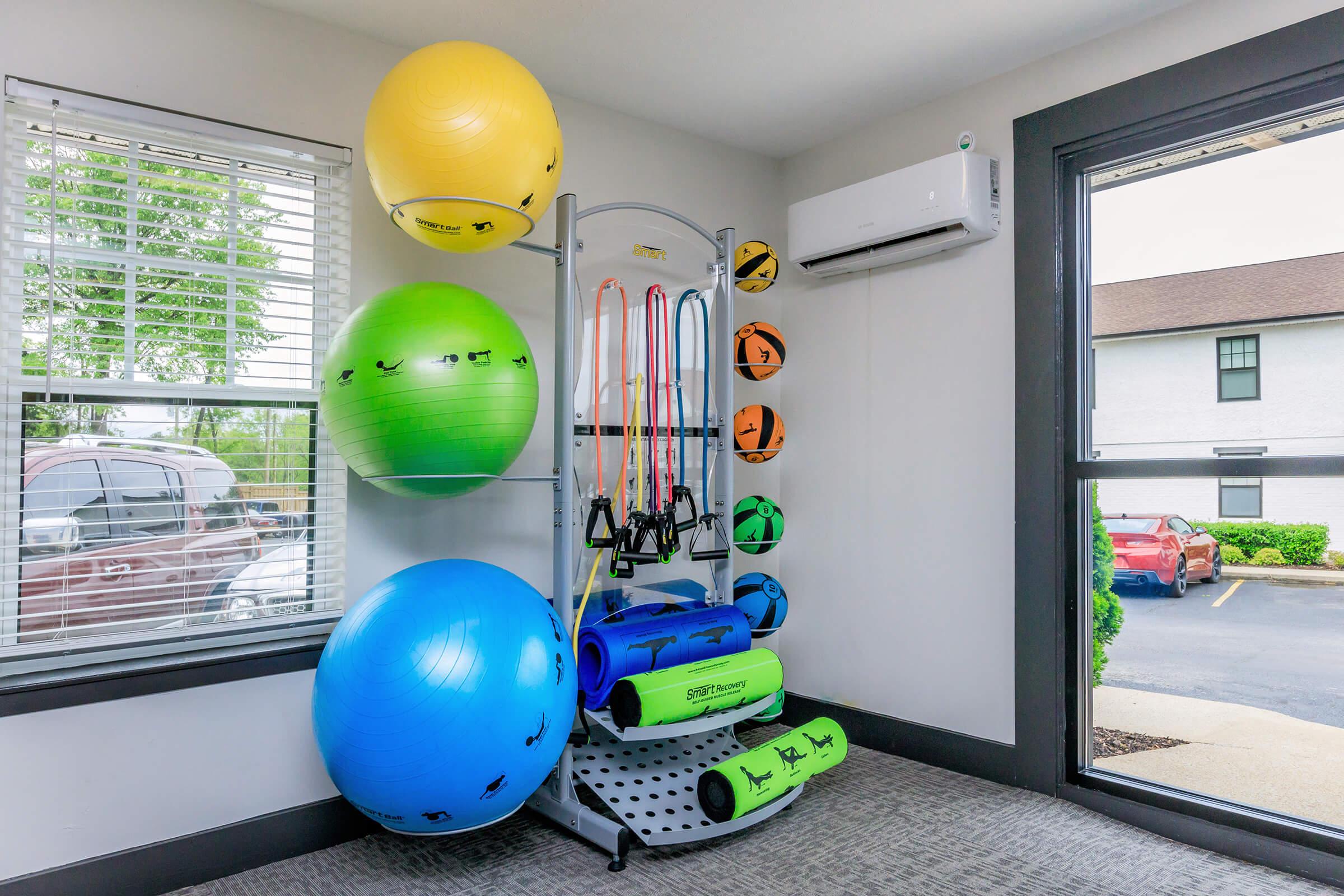A fitness room featuring colorful exercise balls (yellow, green, blue) stacked beside a wall-mounted rack. The rack holds resistance bands and a collection of yoga mats in blue and green. Large windows allow natural light to illuminate the space, with a view of parked cars outside.