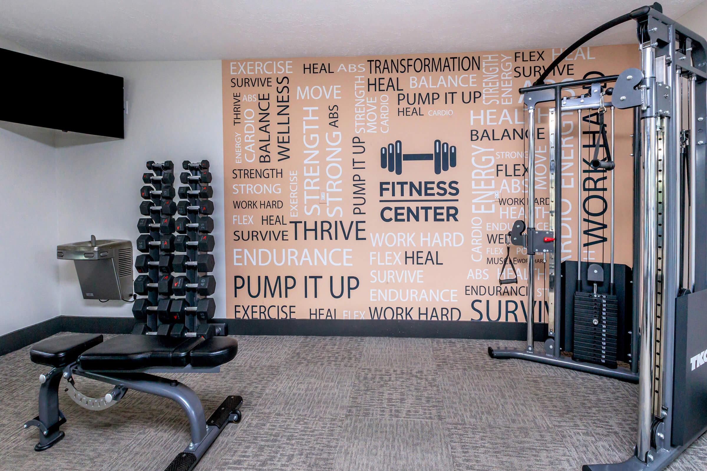 A modern fitness center featuring weight training equipment, including a bench and dumbbells. The wall displays motivational words related to fitness and wellness, such as "strength," "endurance," and "transformation." A water fountain is visible in the background.
