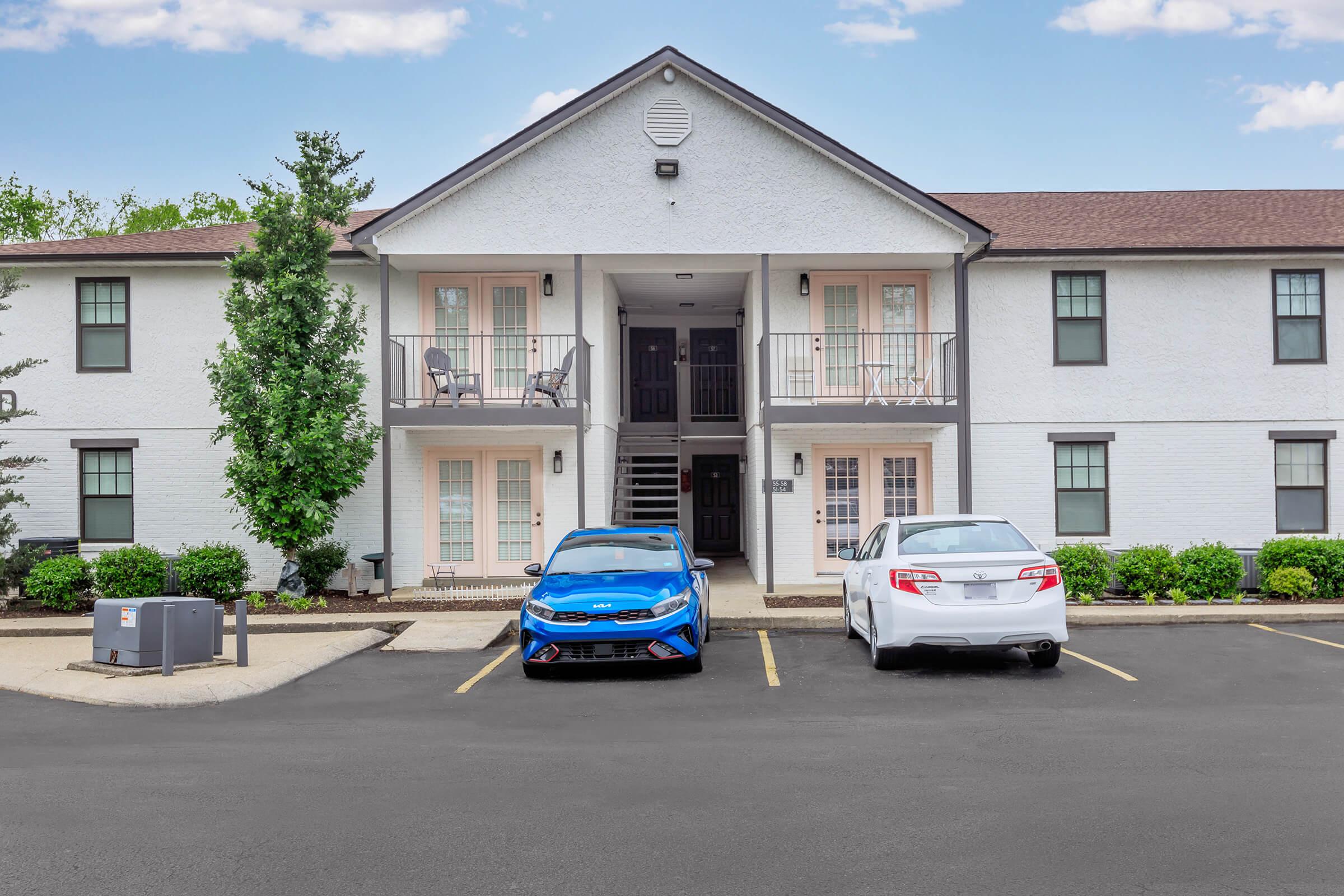 A two-story apartment building with a white exterior. The ground floor features two sets of double doors leading to the entrance, flanked by small balconies. Two parked cars are in front, one blue and one white. Lush green shrubs are planted along the walkway, and a clear blue sky is visible above.