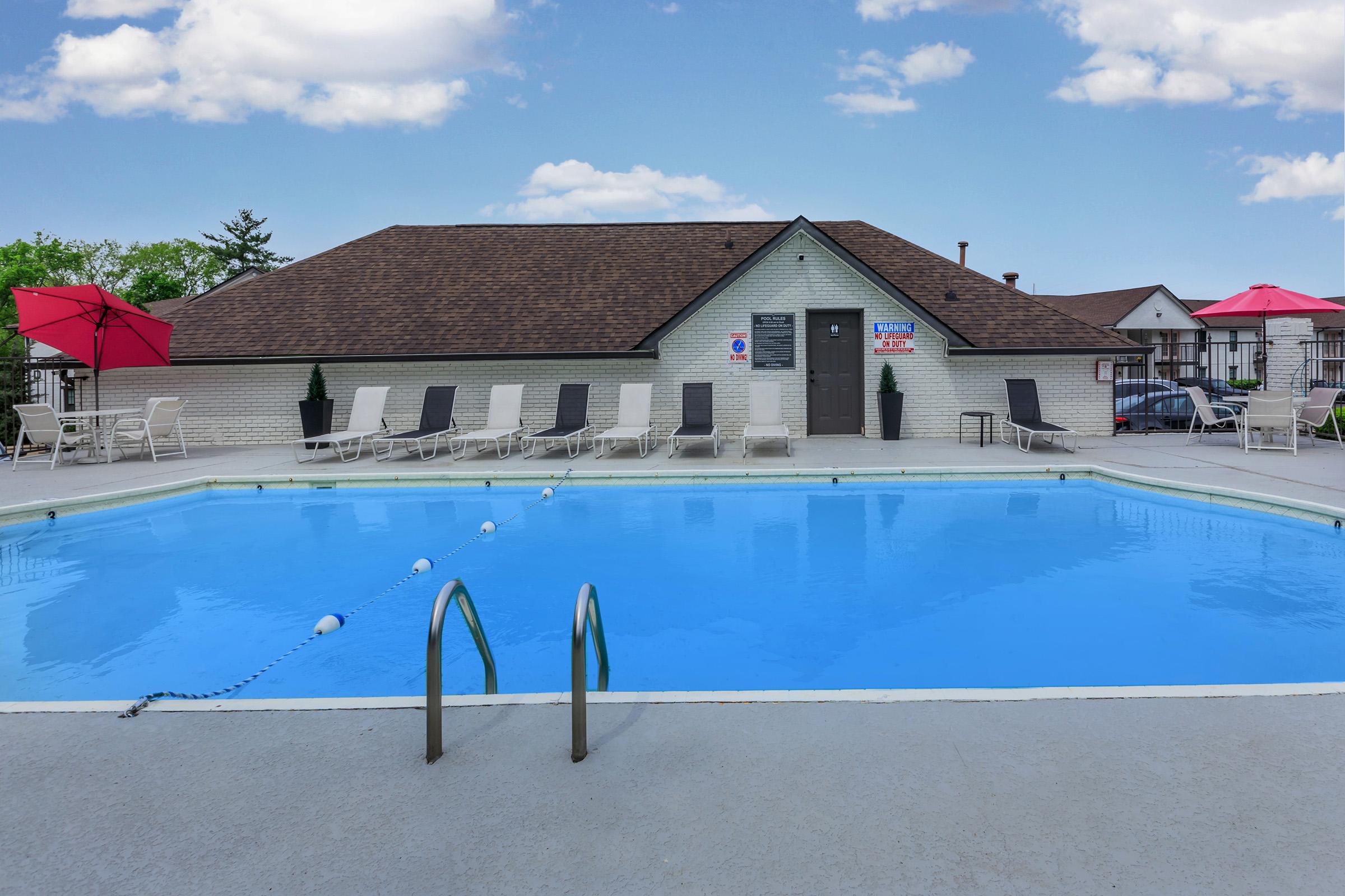 Swimming pool with clear blue water, surrounded by lounge chairs and red umbrellas. A single diving board is visible next to the pool. The background features a building with a brown roof and signs, under a clear blue sky with a few clouds.