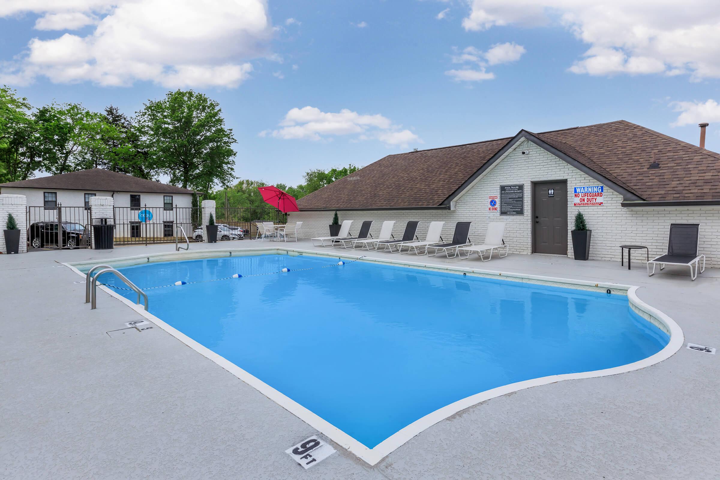 A clear blue swimming pool surrounded by lounge chairs and a shaded area, with a building in the background. The pool deck features a light-colored surface, and there are trees nearby under a partly cloudy sky. The atmosphere is inviting and perfect for relaxation.
