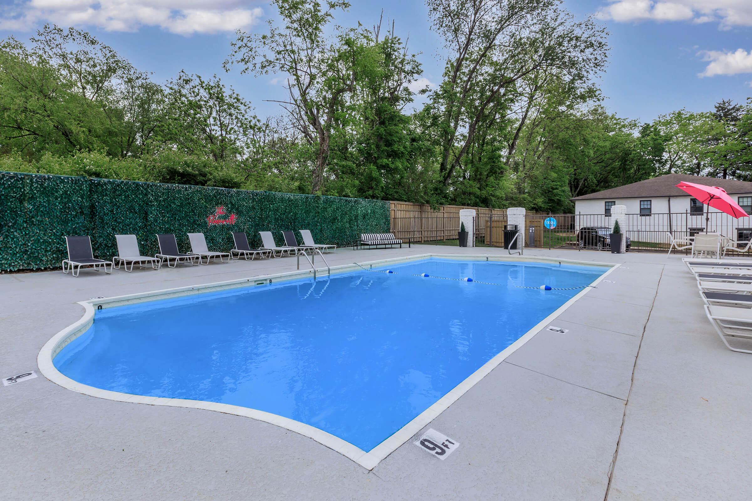 A bright blue swimming pool surrounded by a concrete deck. There are several lounge chairs lined up along one side, and a red umbrella in the corner. Lush green trees and a privacy fence create a serene atmosphere in the background.