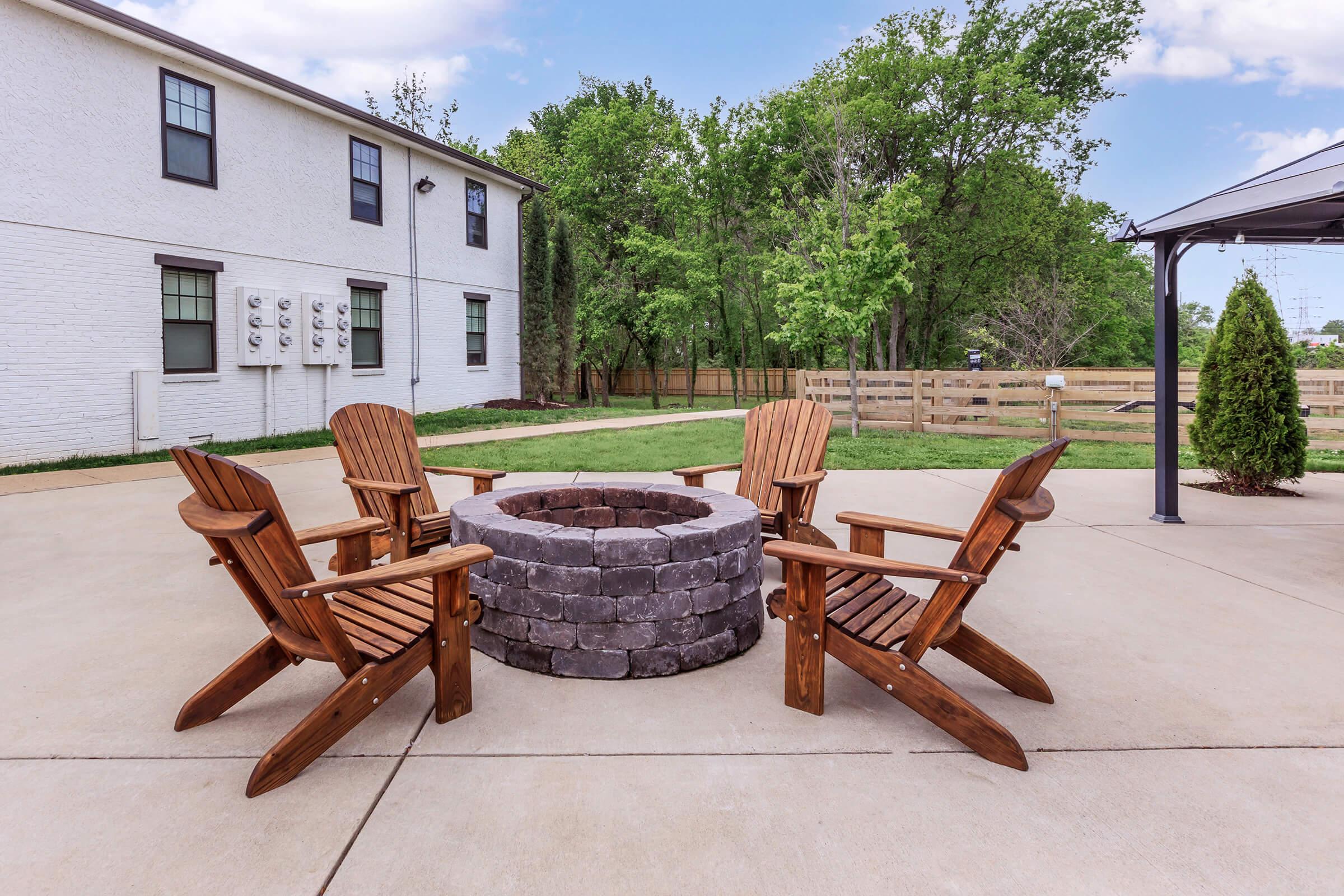 A cozy outdoor fire pit area featuring a circular stone fire pit surrounded by four wooden Adirondack chairs. The setting is surrounded by lush green trees and grass, with a concrete patio in the foreground. In the background, there is a white building and a fenced yard.