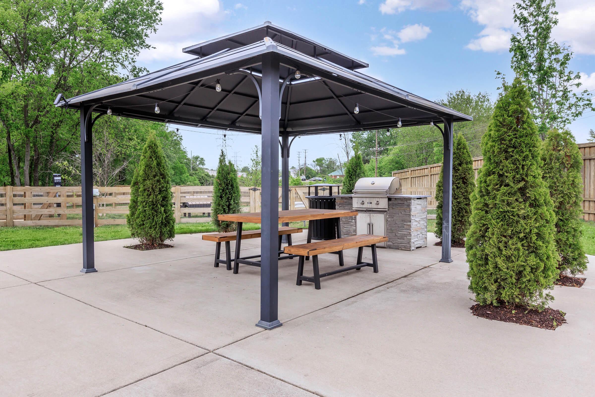 A covered outdoor patio featuring a wooden picnic table, a grill, and landscaped greenery with small trees. The structure has a modern design with a dark metal frame and a tent-like roof, set on a concrete surface, surrounded by a wooden fence and open grass area.