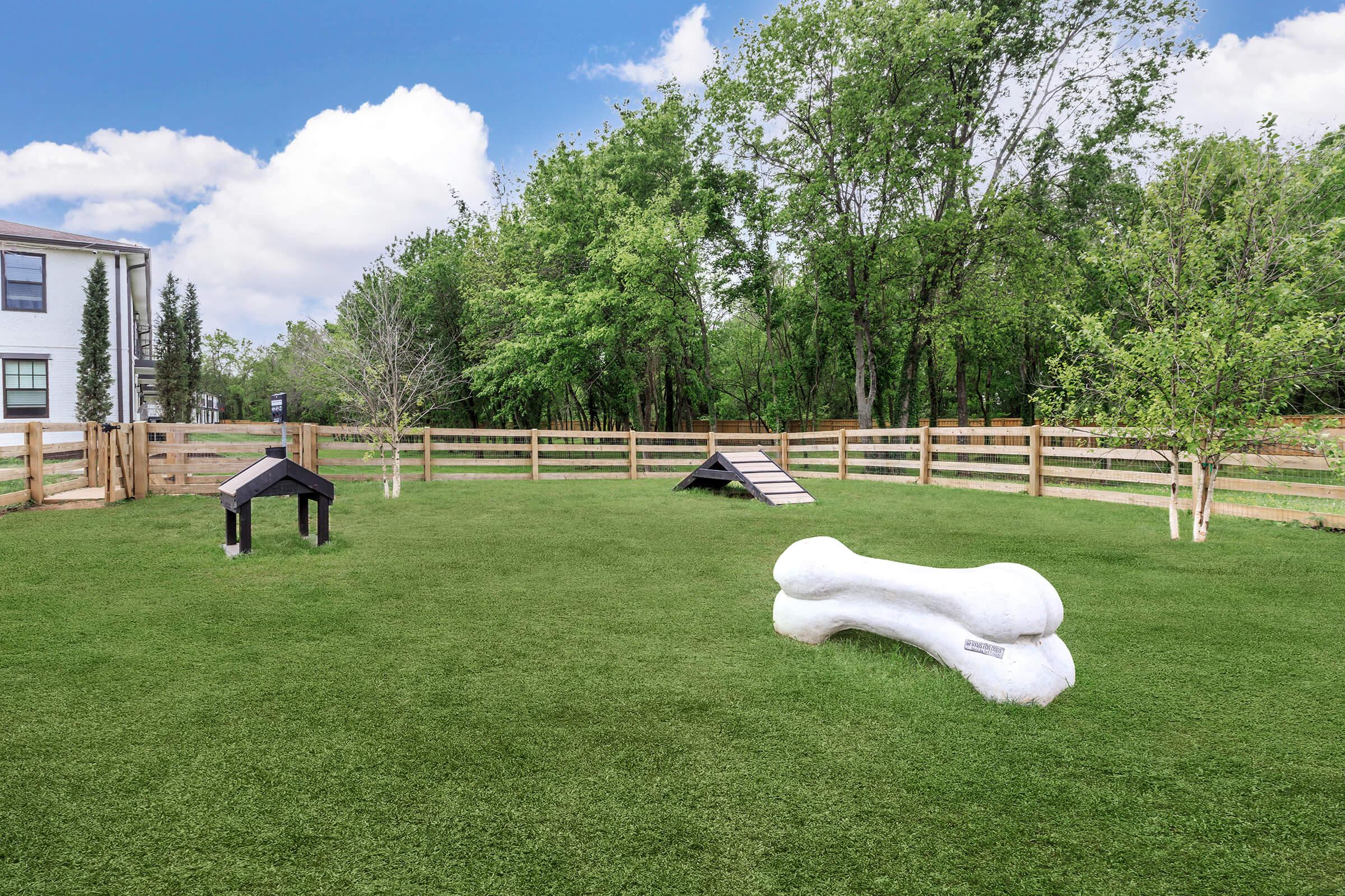 A spacious, well-maintained dog park featuring green grass, a large white dog bone sculpture, a small wooden dog house, and an agility ramp. Surrounding the area are trees and a wooden fence, with a bright blue sky and fluffy clouds in the background.
