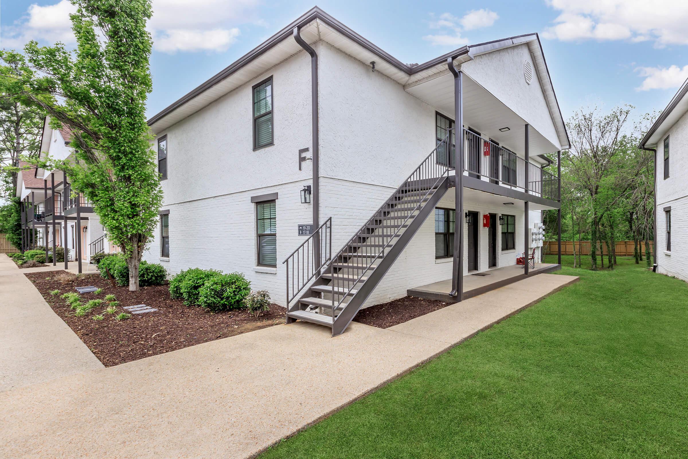 A well-maintained multi-unit residential building with two floors, featuring a gray staircase leading to the upper level. The exterior is painted white with black railings and door accents. Surrounding greenery includes small shrubs and a well-kept lawn, with a pathway leading to the entrance.