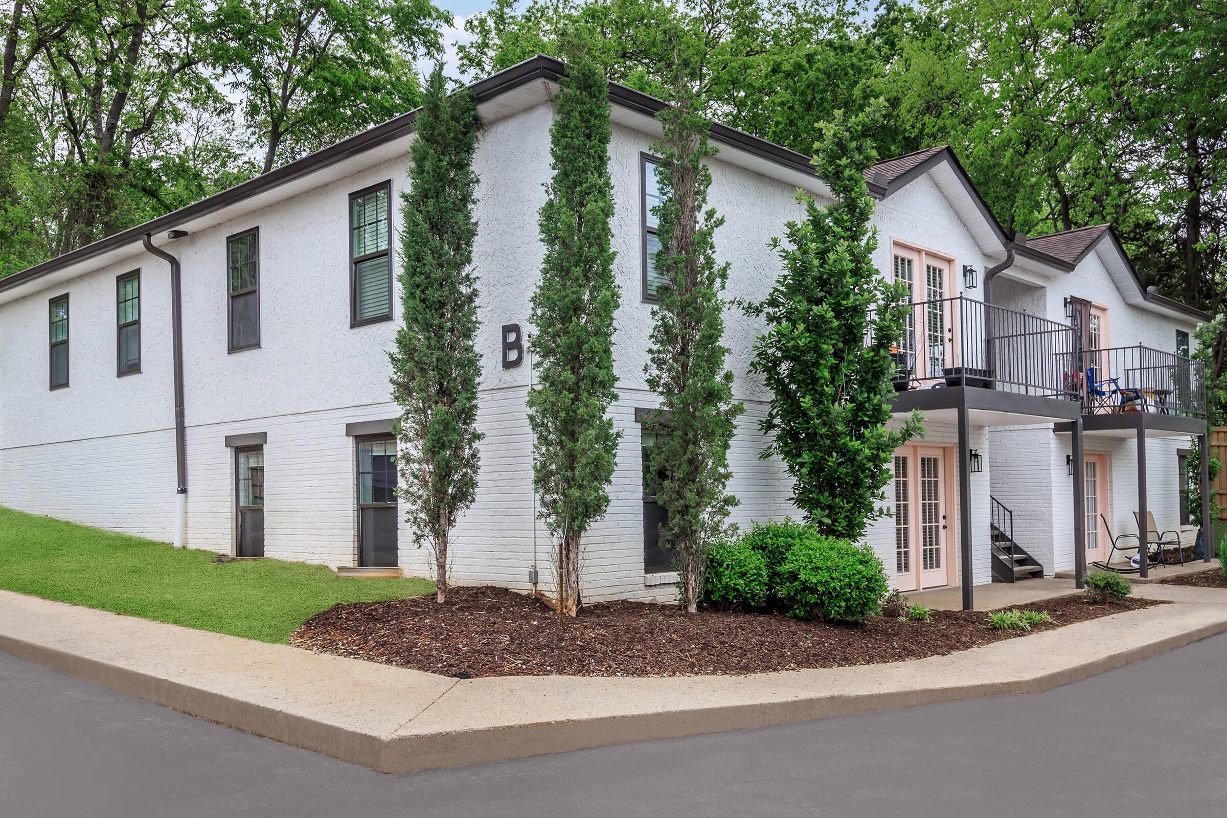 A modern two-story white building with pink accents and large windows, surrounded by green grass and neatly trimmed shrubs. The entrance features a small porch with a railing. Trees line the property, providing a natural setting. A paved pathway curves around the building.
