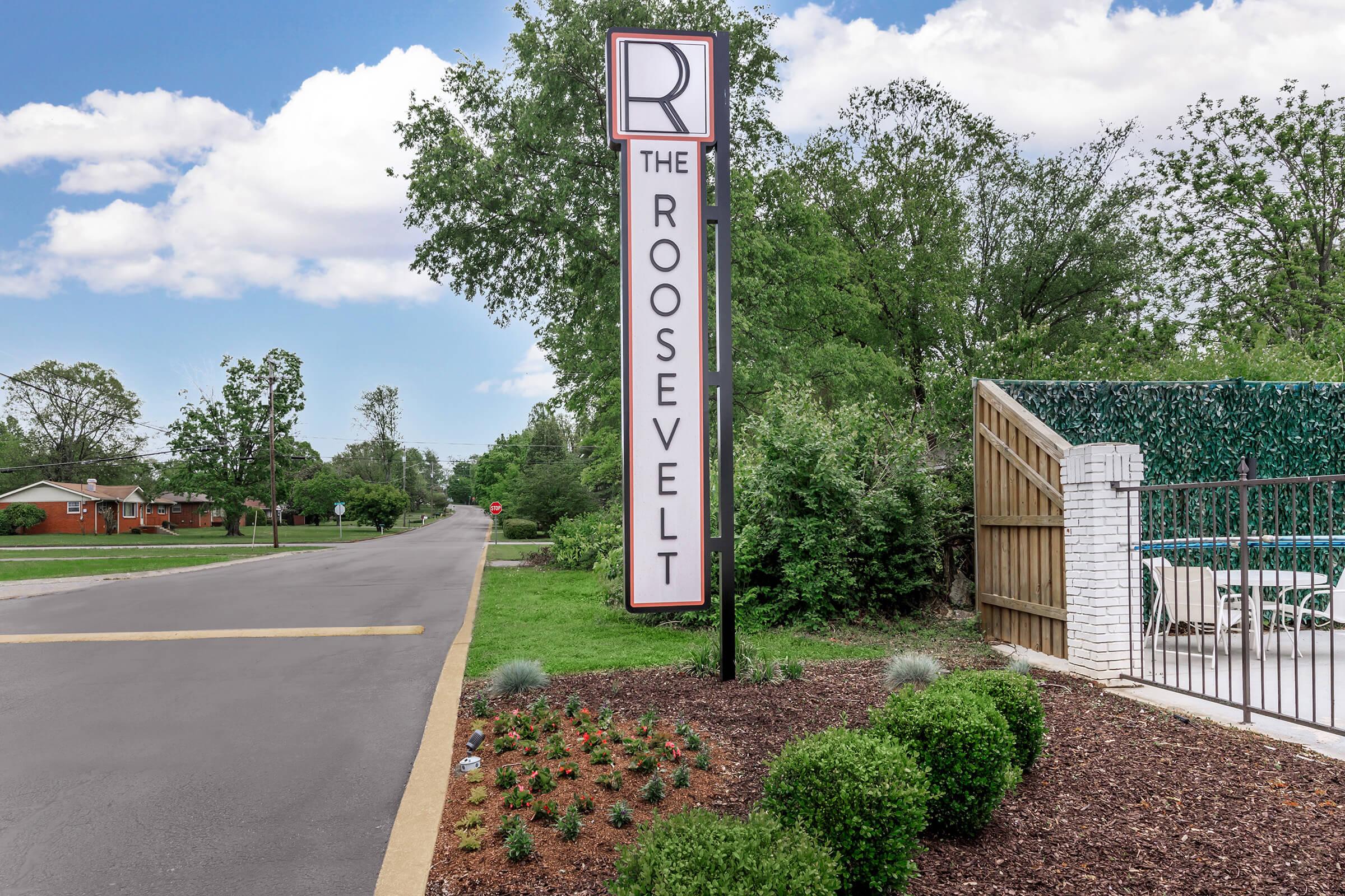 Sign for "The Roosevelt" located along a road, surrounded by greenery. The sign features a large letter "R" at the top. There is a neatly landscaped area with small shrubs in the foreground and a wooden fence partially visible in the background.