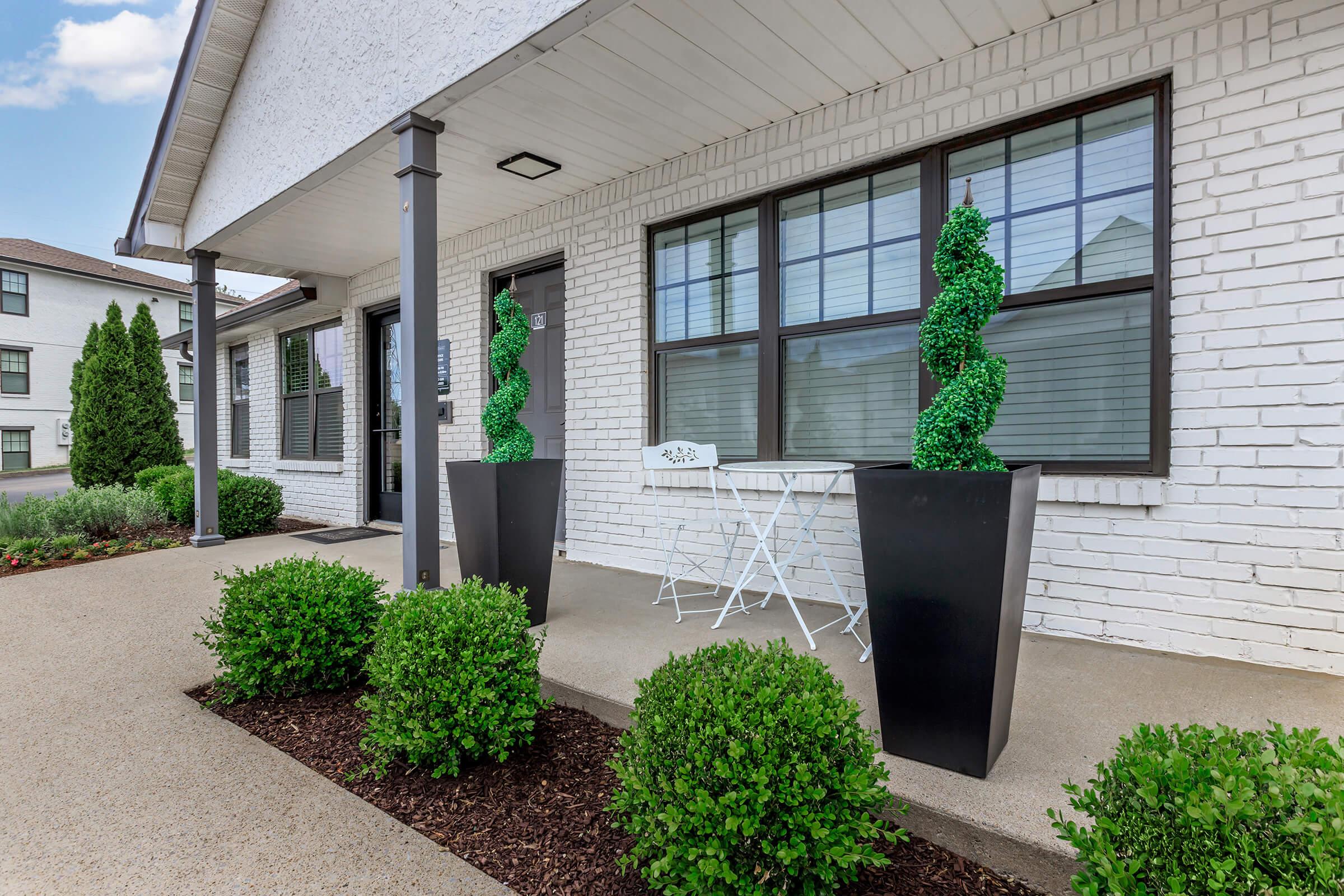 A modern entrance featuring two tall, spiral topiary plants in decorative black planters. A small white table with two chairs is positioned beside the plants. The building has a clean, white brick exterior and large windows, creating a welcoming atmosphere. Lush green shrubs line the pathway.