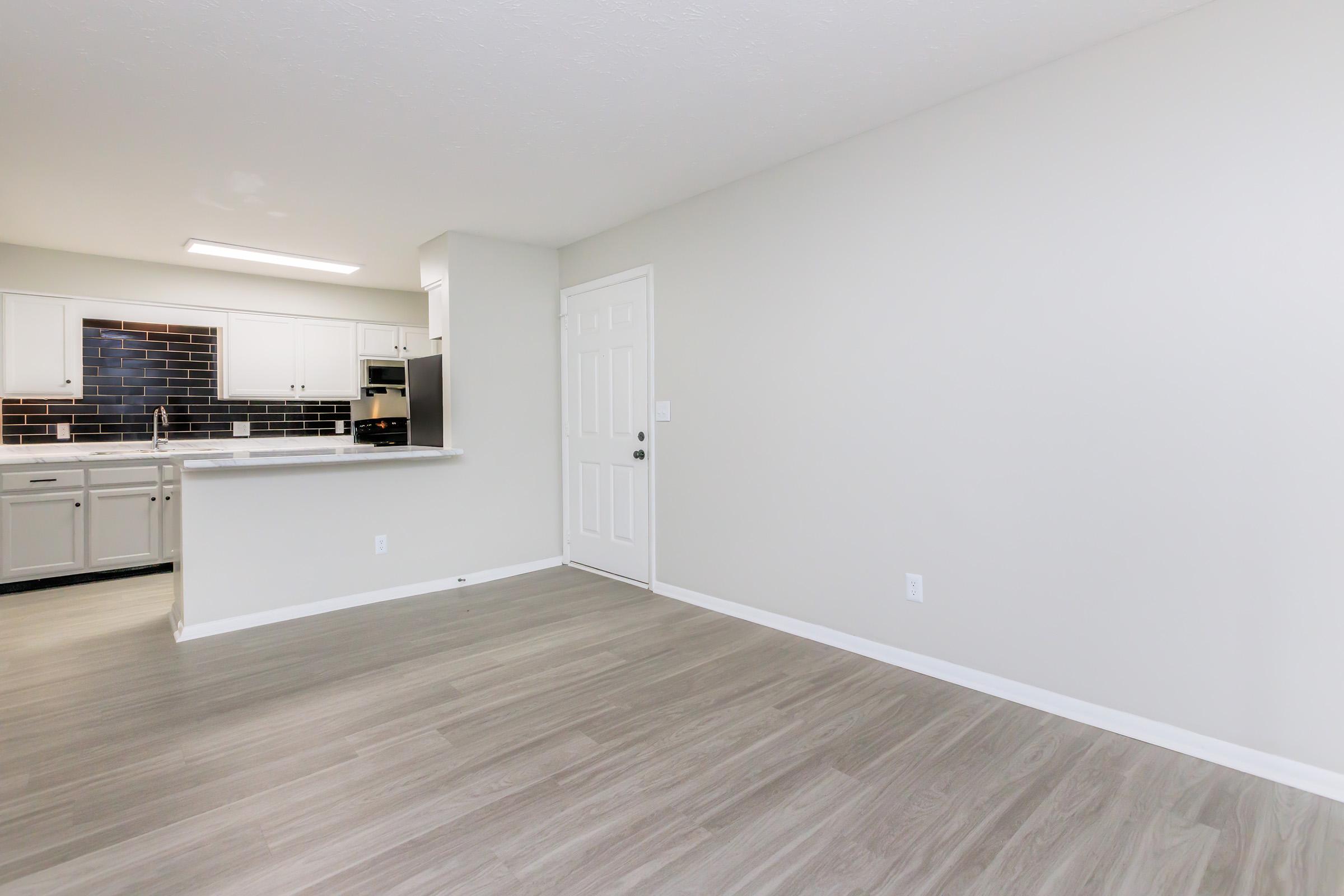 Interior view of a modern, unfurnished living space featuring light gray walls, sleek vinyl flooring, and a small kitchen area with white cabinetry and dark backsplash. The entrance door is visible, along with a window bringing in natural light.