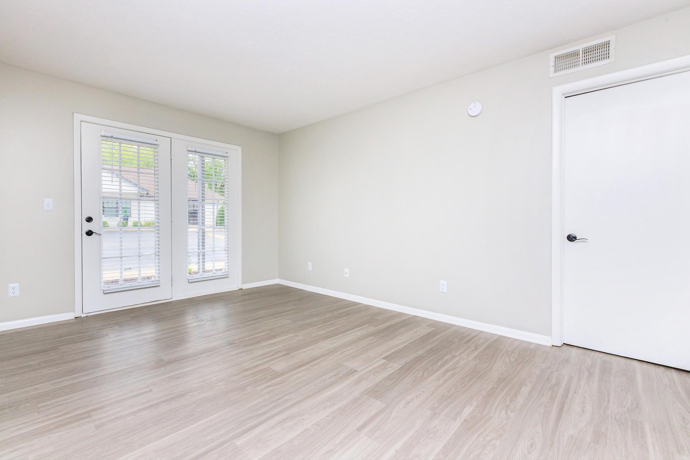 A spacious, empty room with light-colored walls and a light wood floor. Two glass doors with white shutters lead to an outdoor area. A white door on the right side of the image complements the minimalist design, and a small vent is located near the ceiling. The overall atmosphere is bright and inviting.