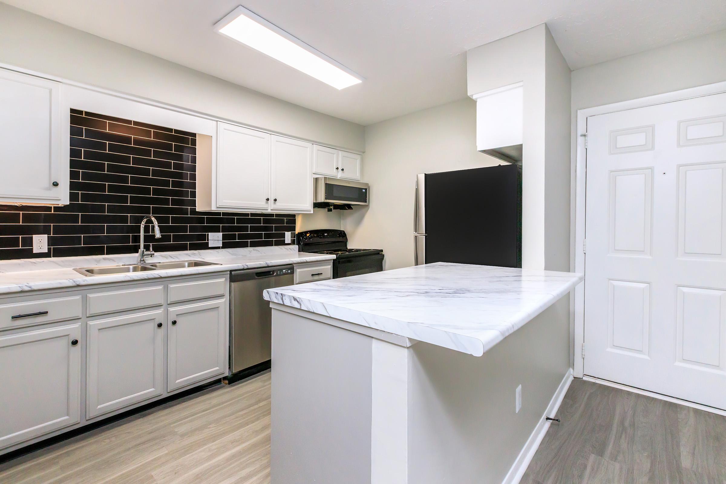 Modern kitchen featuring white cabinets, a black tile backsplash, stainless steel appliances, and a white marble island. The space includes a spacious layout with overhead lighting and a door leading outside, complemented by a light-colored floor.