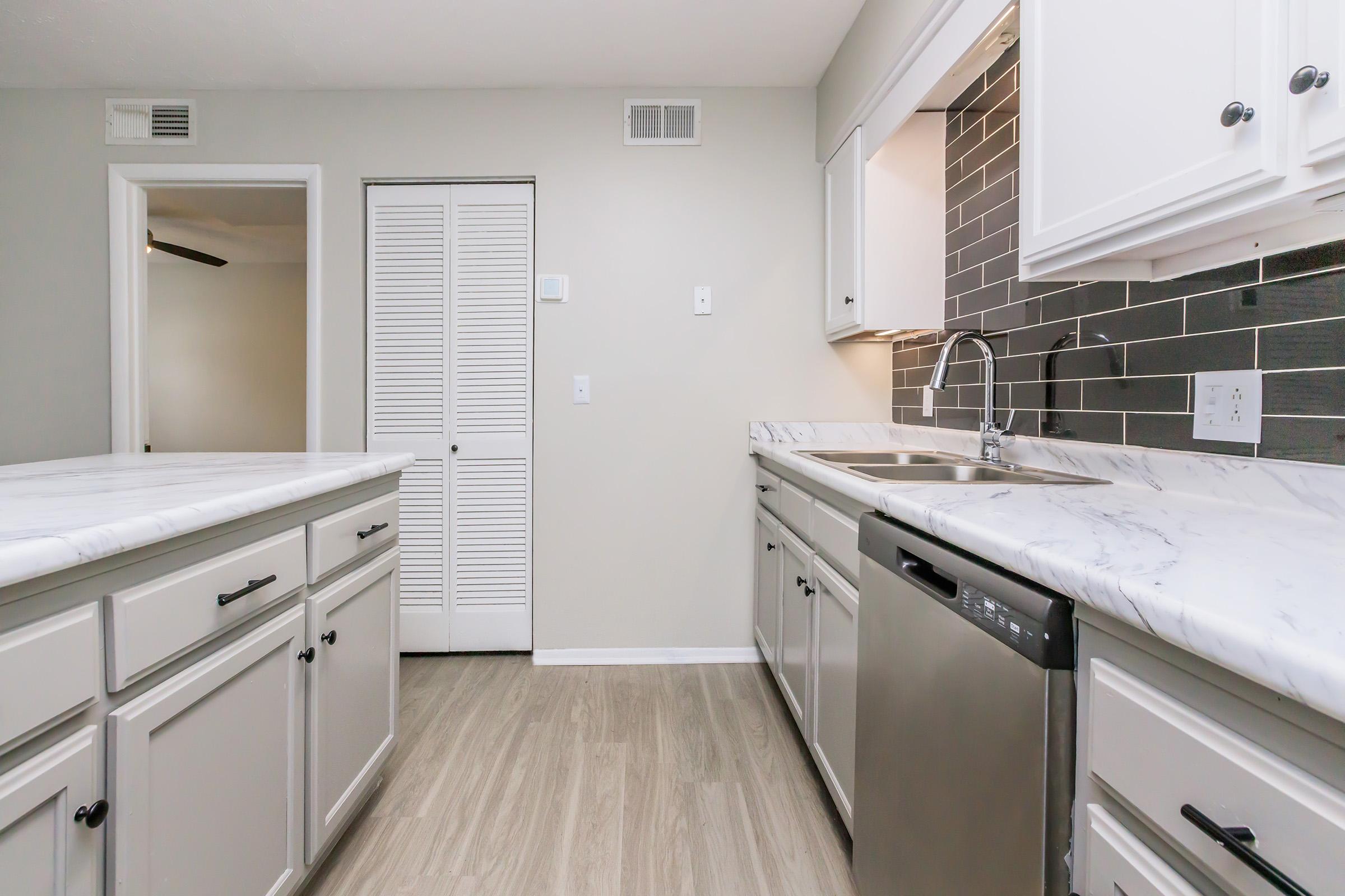 A modern kitchen featuring white cabinetry, a marble countertop, stainless steel sink, and dishwasher. The backsplash is dark gray tiles. There are two kitchen islands and a doorway leading to another room, with a white closet door in the background. The flooring is light wood.