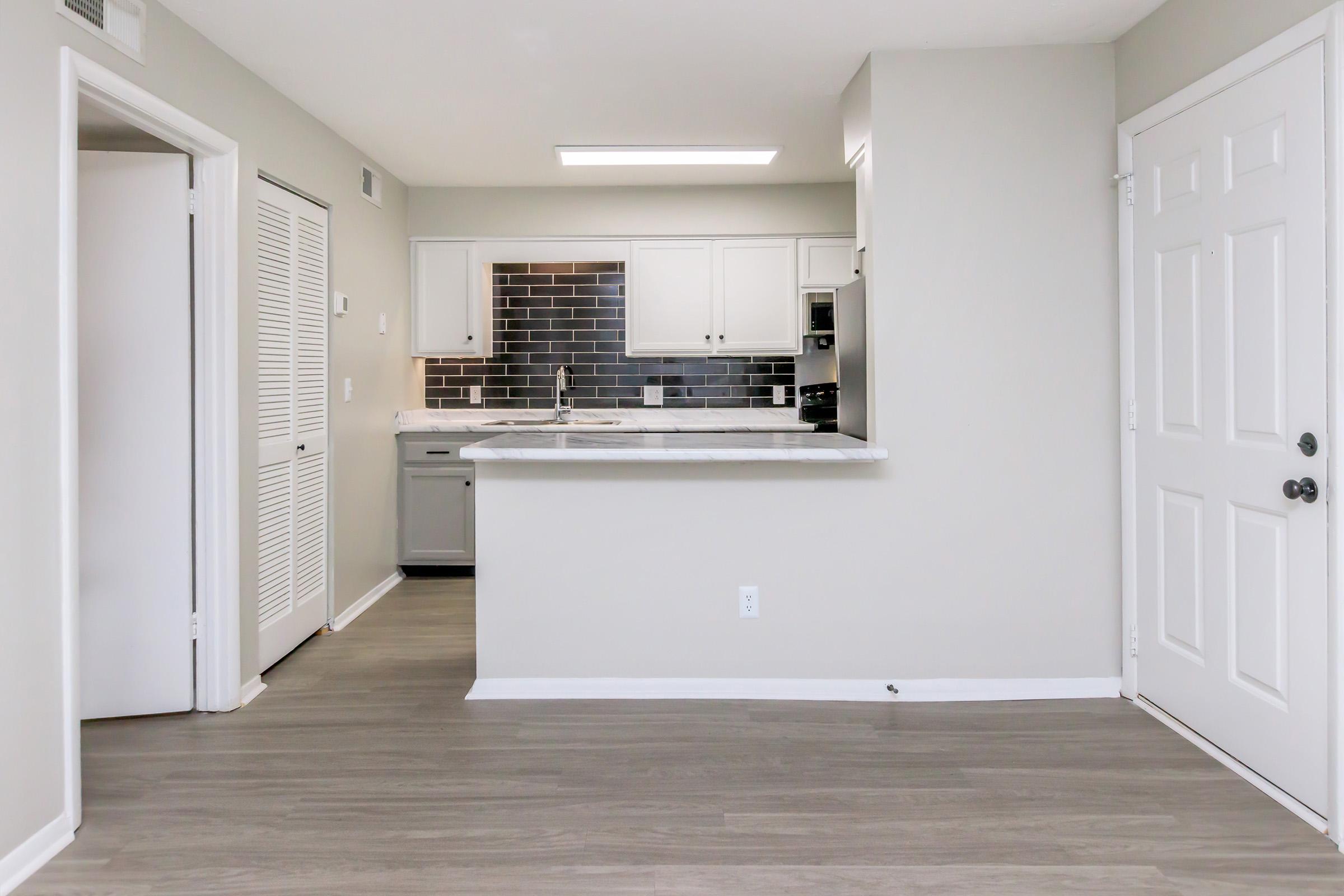 A modern, open-concept kitchen and living space featuring a light gray color scheme. The kitchen includes white cabinets, dark gray tile backsplash, and a marble island. There's a white door on the right and a closet with double doors on the left, with hardwood-style flooring throughout.