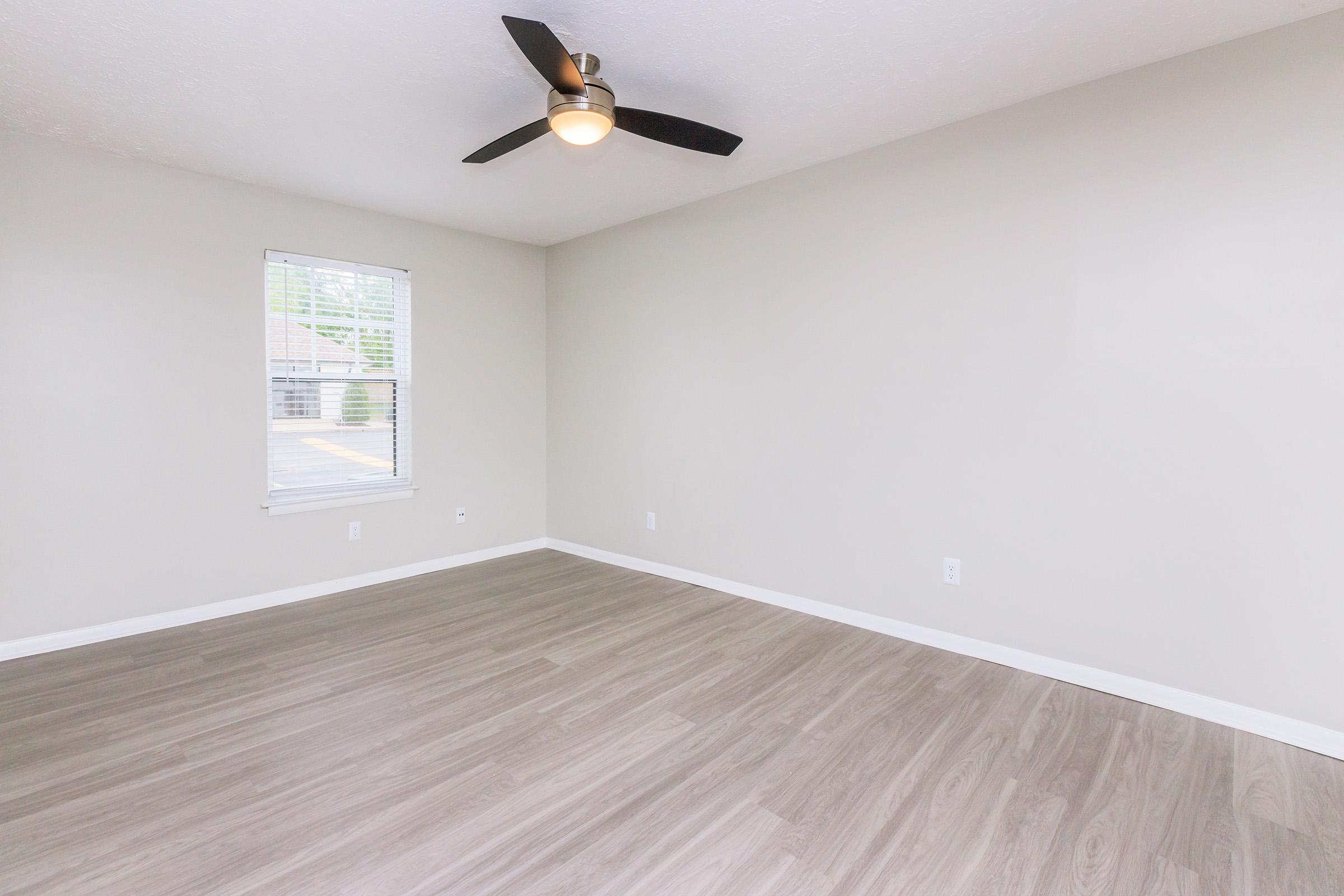 A spacious and empty room featuring light-colored walls, a ceiling fan, and a large window with blinds. The floor is covered in light wood-like laminate, creating a bright and inviting atmosphere.