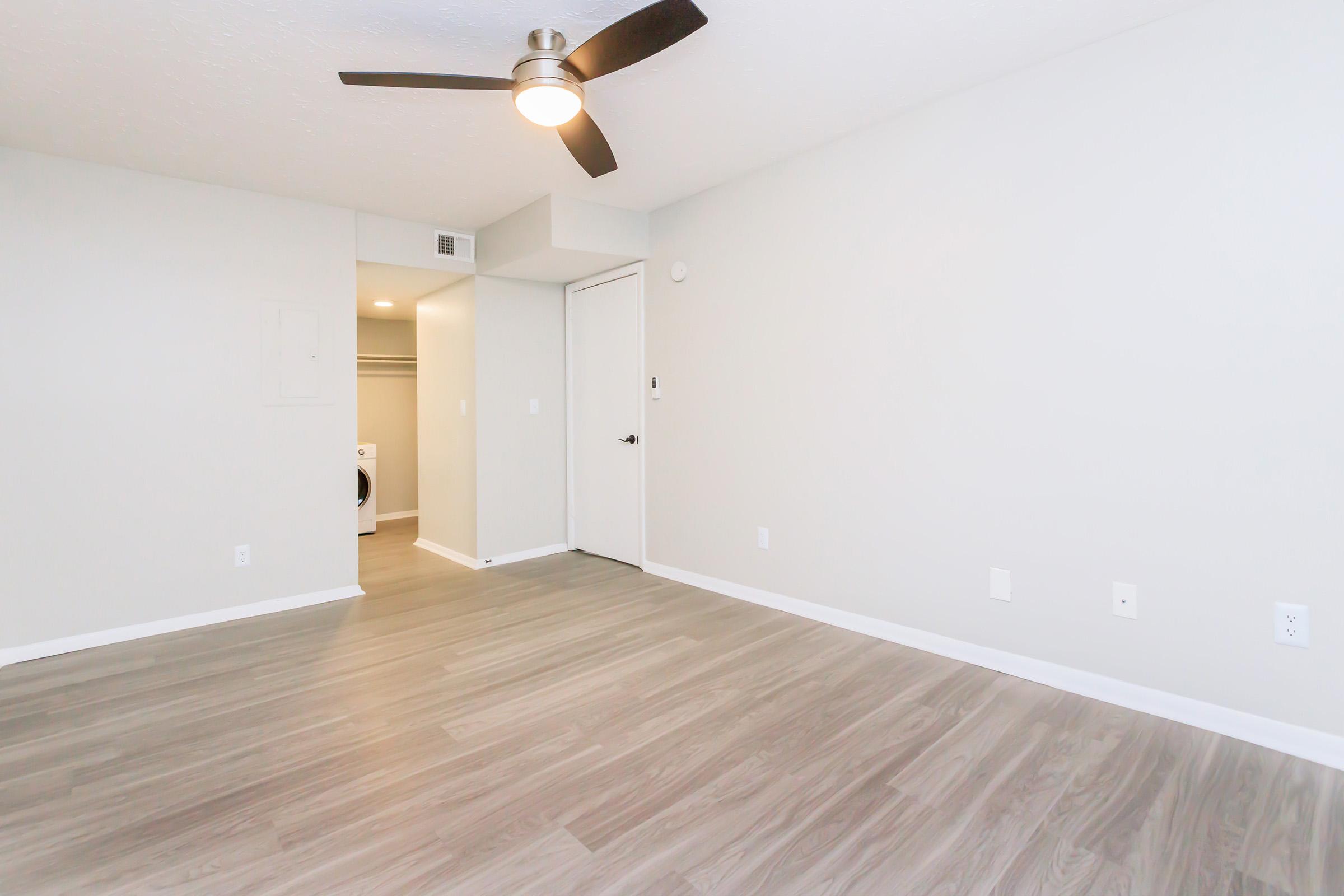 A well-lit room with light gray walls and a ceiling fan. The floor is made of light wood laminate. A closet is visible on the left with a white door. The room has minimal furnishings, creating an open and spacious feel.