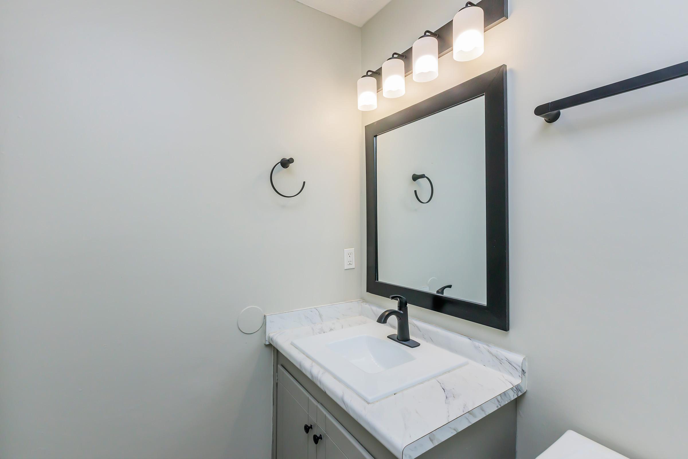 A modern bathroom featuring a vanity with a marble countertop and a rectangular sink. Above the sink, there’s a large wall mirror framed in black, flanked by four round light fixtures. A towel holder is mounted on the wall, and the color scheme is light gray, creating a clean and contemporary look.