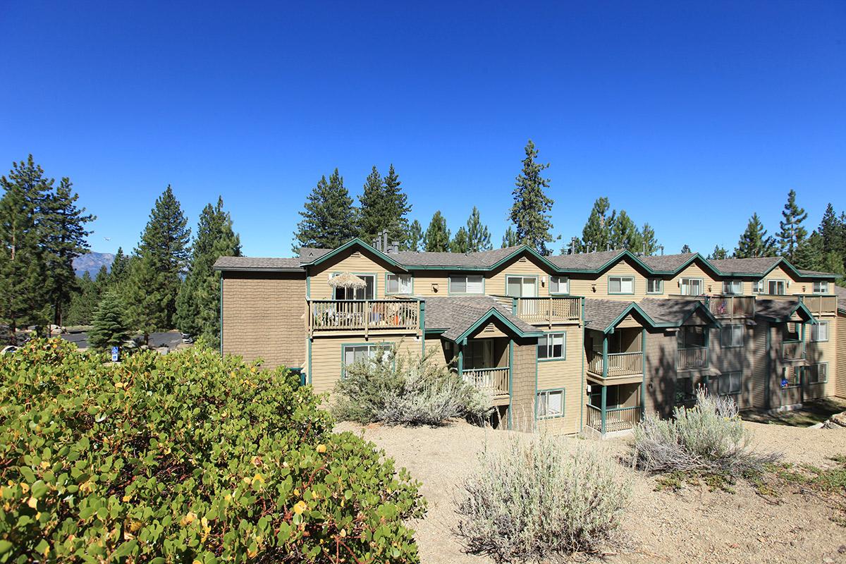 A multi-story brick building surrounded by greenery and trees under a blue sky. The structure features balconies and is situated in a natural landscape, suggesting a residential or vacation property in a mountainous or forested area.