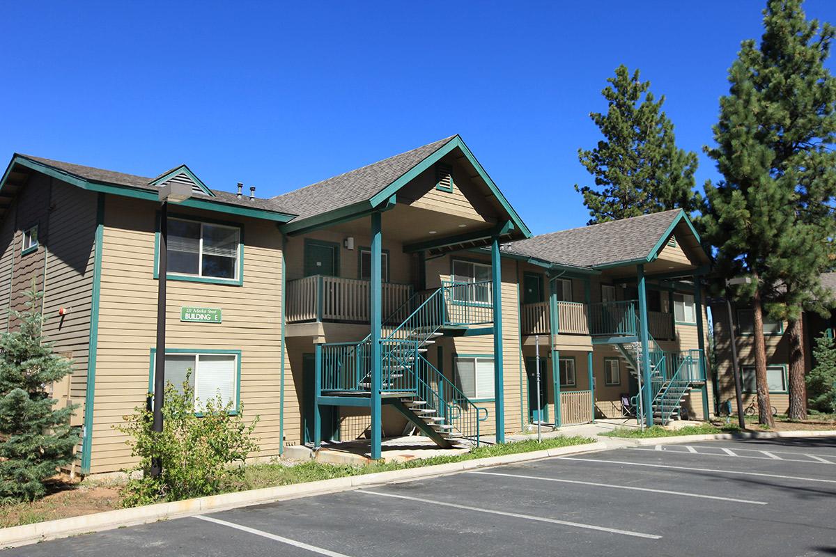 Two-story wooden apartment building with green trim, featuring multiple entrances and a central staircase. Surrounding the building are small trees and parking spaces. The sky is clear and blue, suggesting a sunny day.