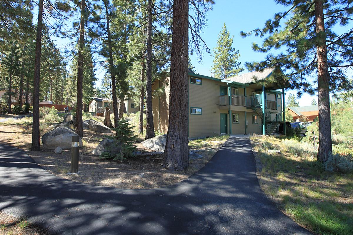 A two-story wooden building surrounded by tall pine trees and natural landscaping. A pathway leads to the entrance, with large rocks scattered around. Clear blue sky above and other structures visible in the background, suggesting a serene, forested environment.
