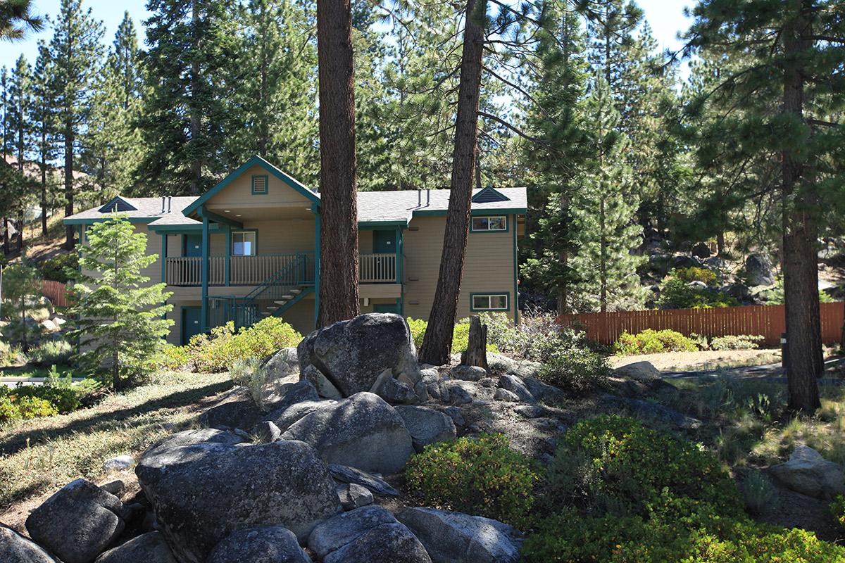 A two-story house surrounded by tall trees and large rocks. The home features a light-colored exterior with teal accents. A wooden fence is visible in the background, and the landscape includes patches of greenery and shrubs. Sunlight filters through the trees, creating a serene, natural setting.