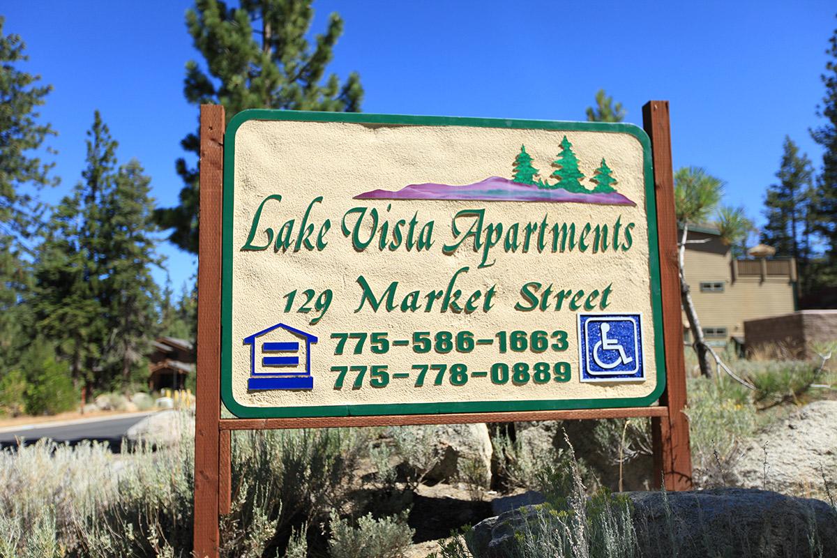 A sign for Lake Vista Apartments displaying the address "129 Market Street" along with phone numbers "775-586-1663" and "775-778-0889." The sign features a nature-themed design with trees and mountains in the background, indicating accessible options with a wheelchair symbol.