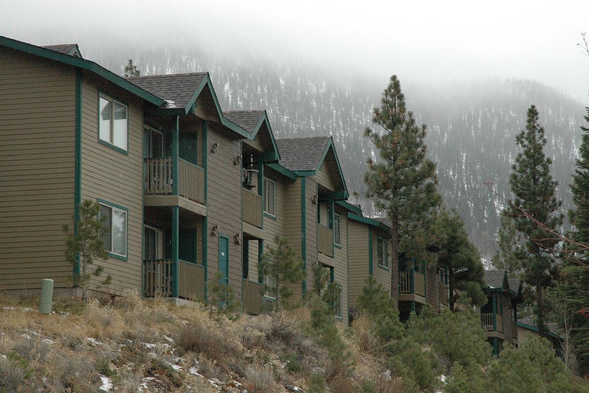 A row of two-story townhouses in a mountainous area, with a misty background featuring snow-capped peaks. The buildings have green trim and balconies, surrounded by pine trees and a grassy foreground. The scene conveys a serene, nature-filled environment.