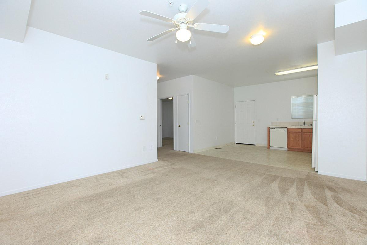 Interior of a spacious, empty living room featuring light beige carpet, white walls, and a ceiling fan. A doorway leads to a kitchen with wooden cabinets and appliances. Natural light enters the room, creating a bright atmosphere. The layout is open, providing a sense of space and versatility for furniture arrangement.
