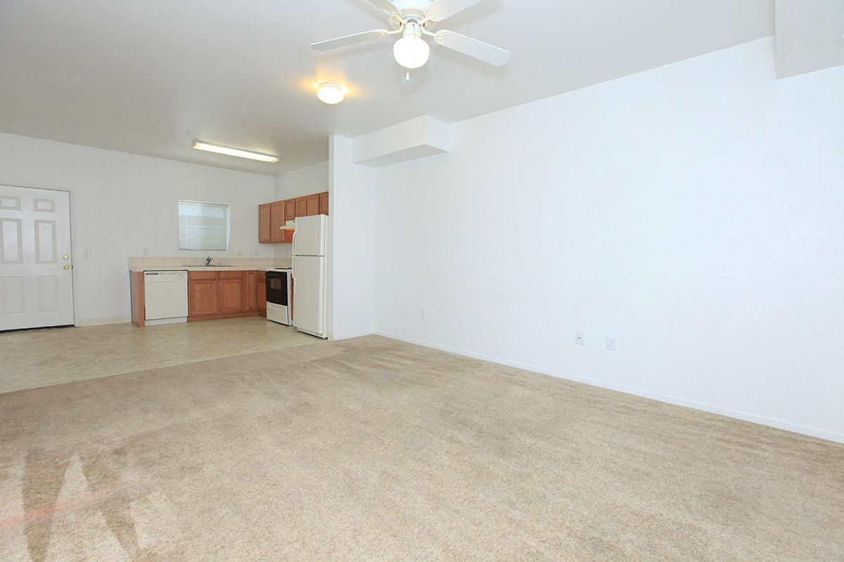 A spacious, empty apartment interior featuring a light-colored carpet, white walls, and a ceiling fan. The kitchen area includes wooden cabinets, a refrigerator, and a dishwasher, while the living area is well-lit with natural light coming from a window.