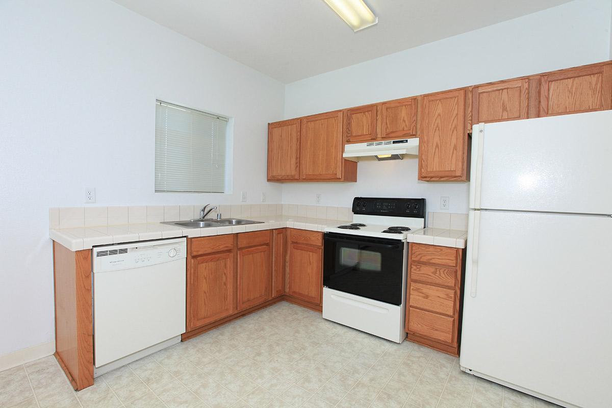 A clean, modern kitchen featuring light-colored walls and laminate flooring. It includes wooden cabinetry, a double sink, a dishwasher, an electric stove, and a white refrigerator. A window lets in natural light, and the countertops are tiled.