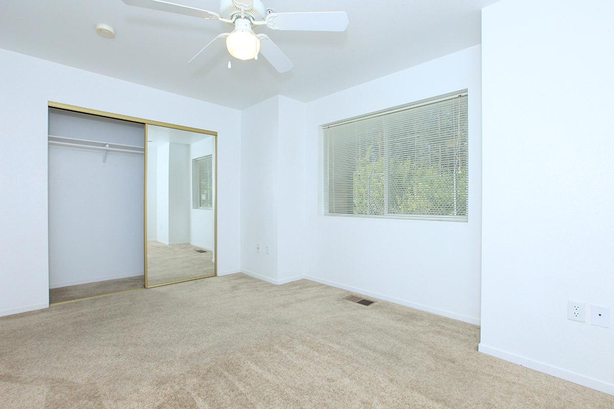 A sparsely furnished room with beige carpet, a ceiling fan, and a window with blinds. A mirrored closet with gold trim is visible on the left. The walls are white, creating a bright and open atmosphere.