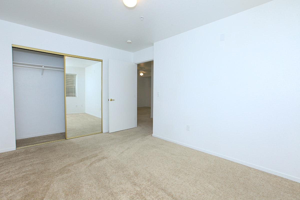 Empty room with light-colored walls and beige carpet. A closet with mirrored sliding doors is visible on the left, while a closed door leads to another room on the right. Natural light comes in from a window, creating a bright and spacious feel.
