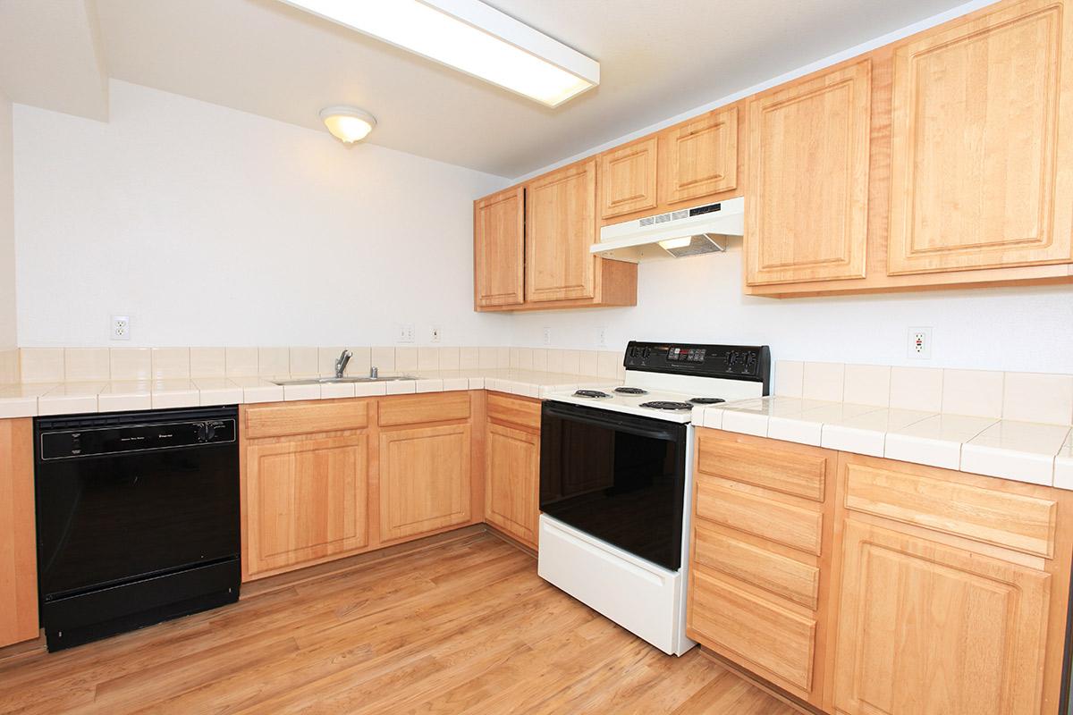 A modern kitchen featuring light wood cabinets, a white stove and microwave, a black dishwasher, and a tiled countertop. The flooring is light wood, and the walls are painted a soft white. The layout includes a sink and ample counter space, creating a functional cooking area.