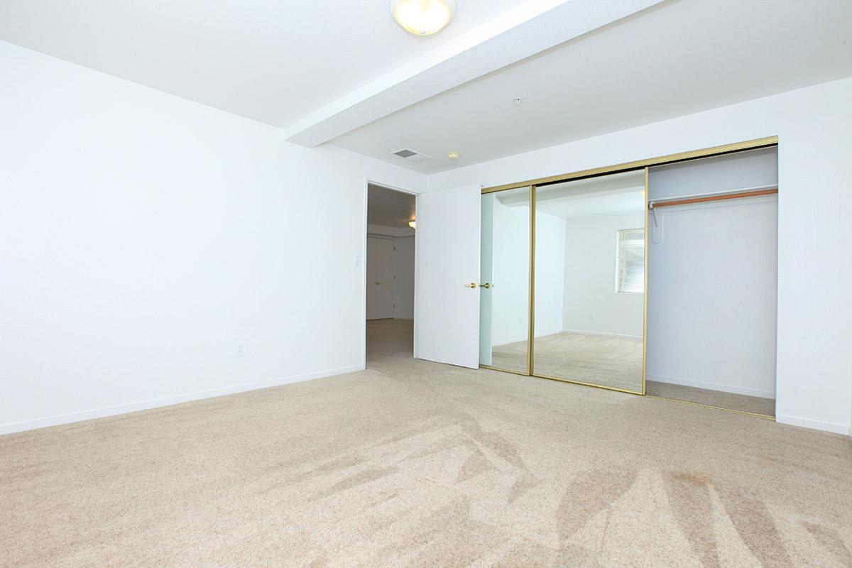 A spacious, empty room with beige carpeting and white walls. There is a large mirrored closet with sliding doors on one side and a ceiling light fixture. The room appears bright and well-lit, with a window visible in the background, casting soft shadows on the carpeted floor.