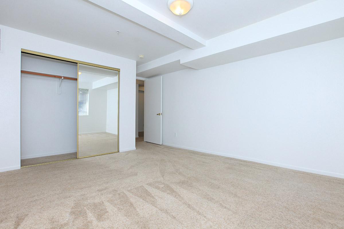 A spacious, empty room with light-colored carpet and white walls. There is a mirrored closet door on one side, and a door leading to another room is visible. The room has a ceiling light fixture and a small window that allows natural light to enter.