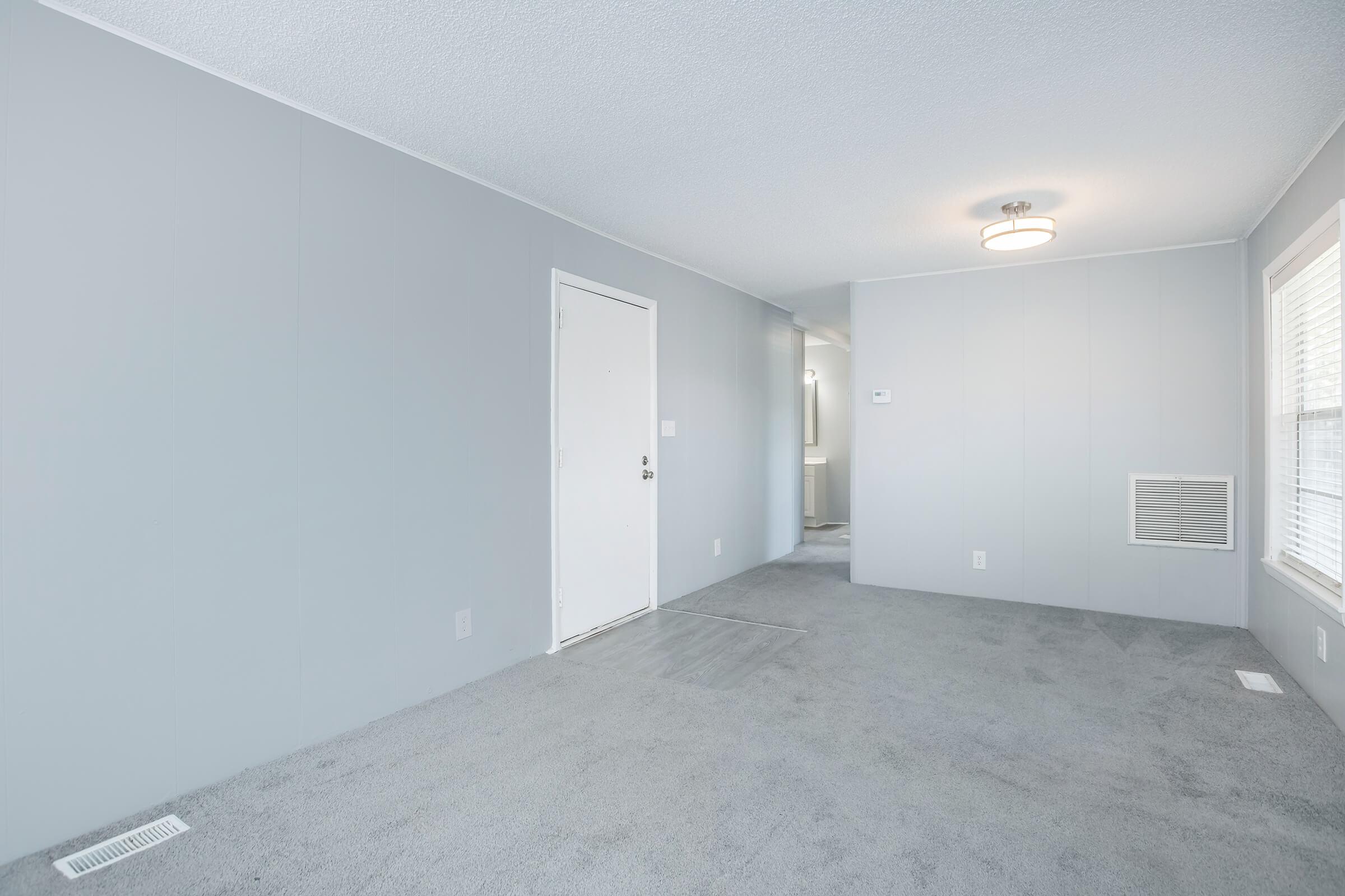 Interior of a light gray room with carpeted flooring. A door is visible on the left side, and there's a vent near the floor. The walls are plain gray, and the room is well-lit with a ceiling light. A small window with blinds is on the right side, but no furnishings are present.