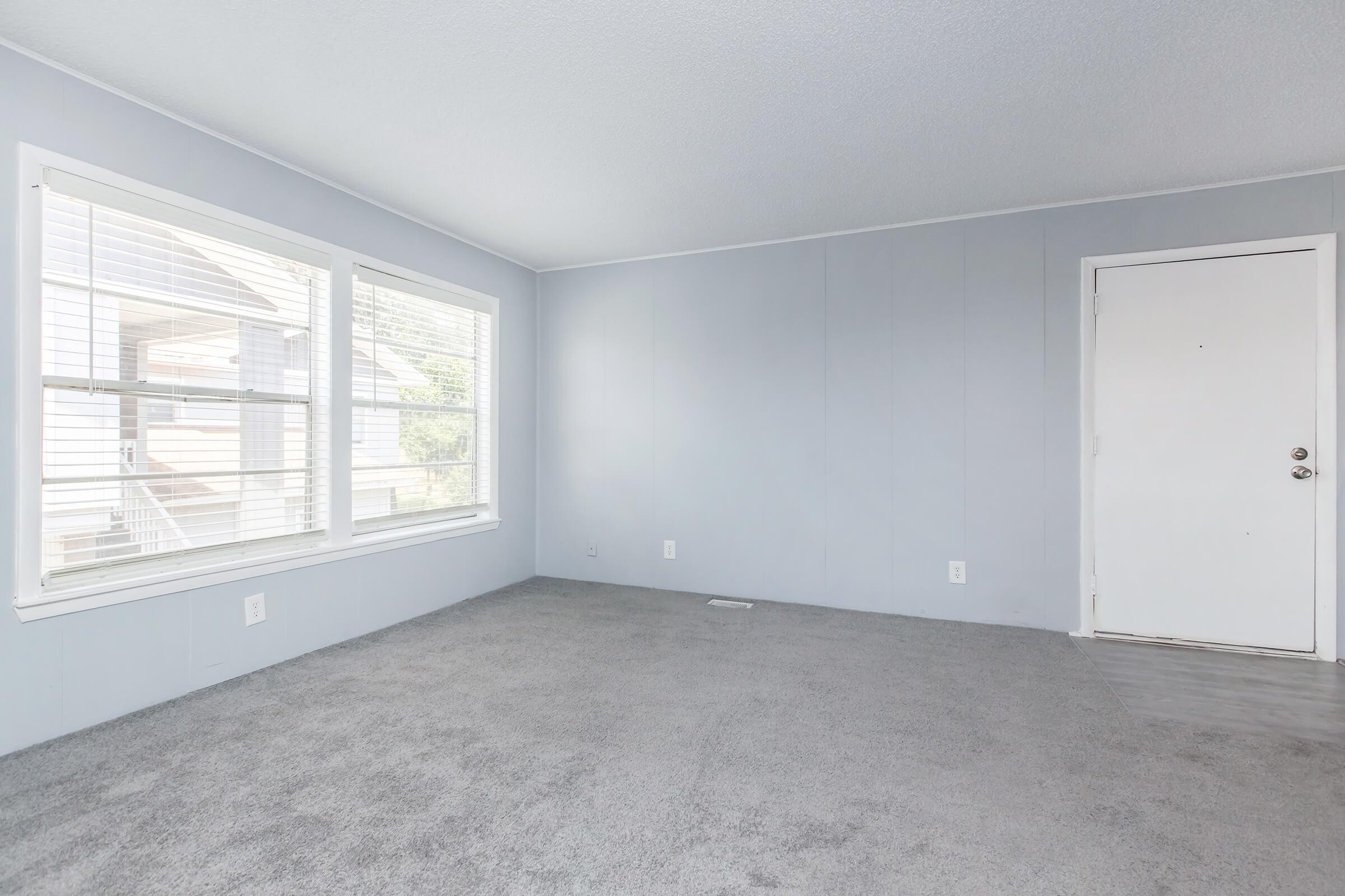 Empty room with light gray walls, carpeted floor, and large windows allowing natural light. There's a white door on one side of the room. The overall atmosphere is bright and spacious, suitable for various uses.