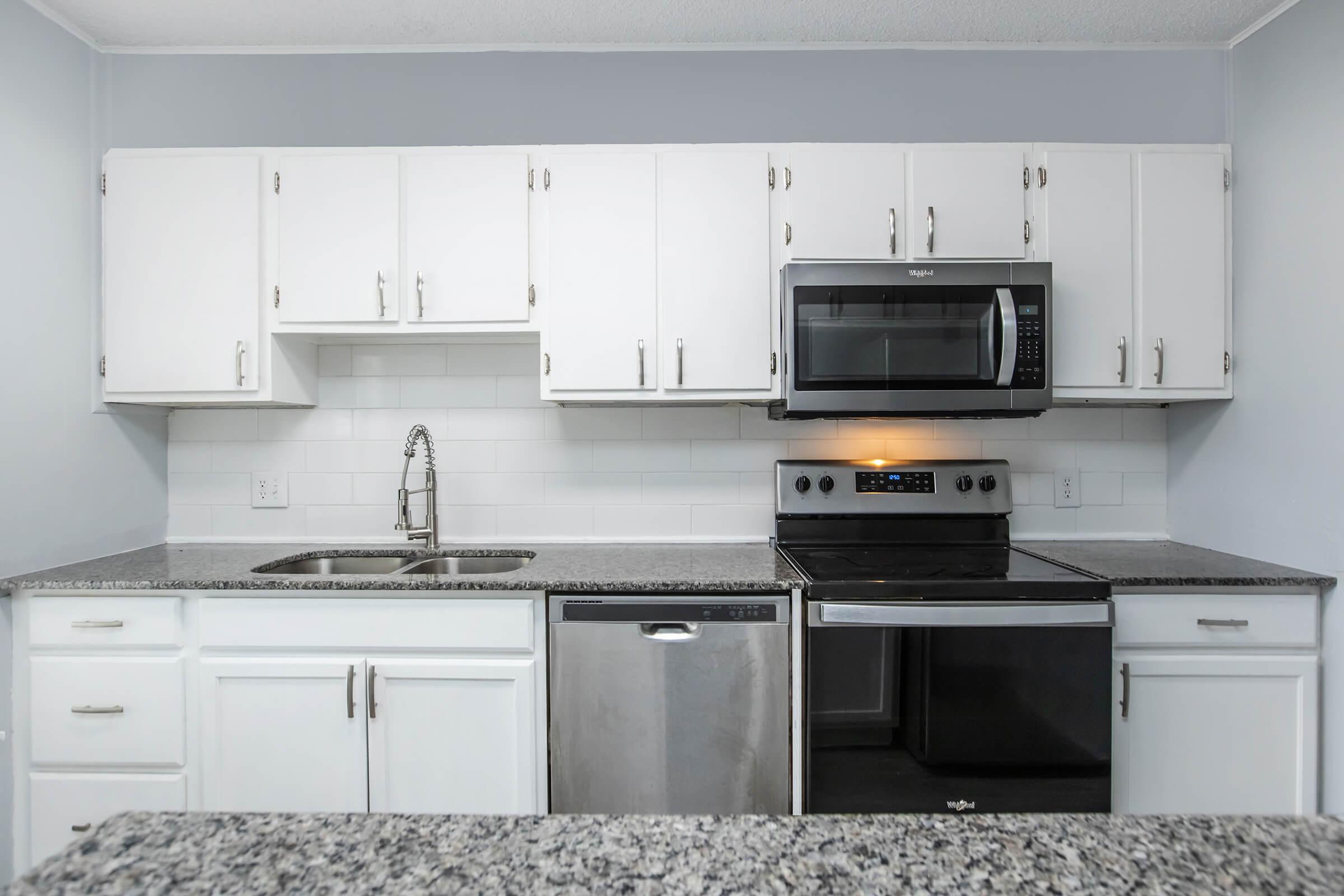 Modern kitchen featuring white cabinets, a stainless steel sink, microwave, and stove, with a granite countertop. A dishwasher is situated beside the sink, and the walls are painted light blue, creating a clean and spacious appearance.