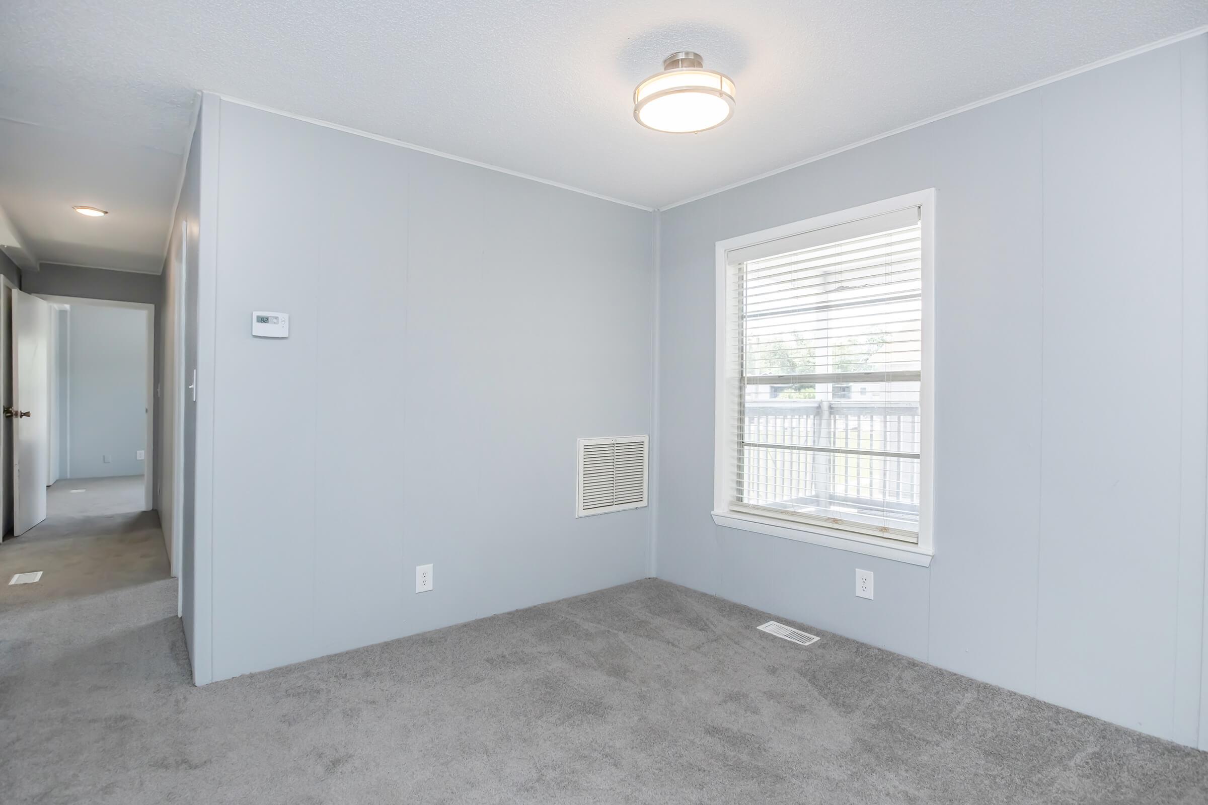 A well-lit interior room with light gray walls and carpet. A large window with white blinds lets in natural light. There's a light fixture on the ceiling and an air vent on the wall. The space appears empty and ready for furniture, with a door visible in the background leading to another area.