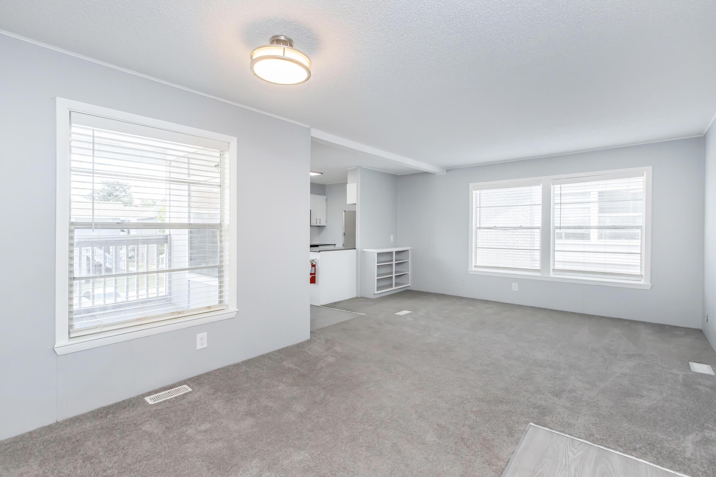 A spacious living room with light gray walls and carpet, featuring two windows that let in natural light. A ceiling light fixture is visible, and an open area leads to a kitchen or dining space in the background with modern cabinetry. The overall atmosphere is bright and inviting.
